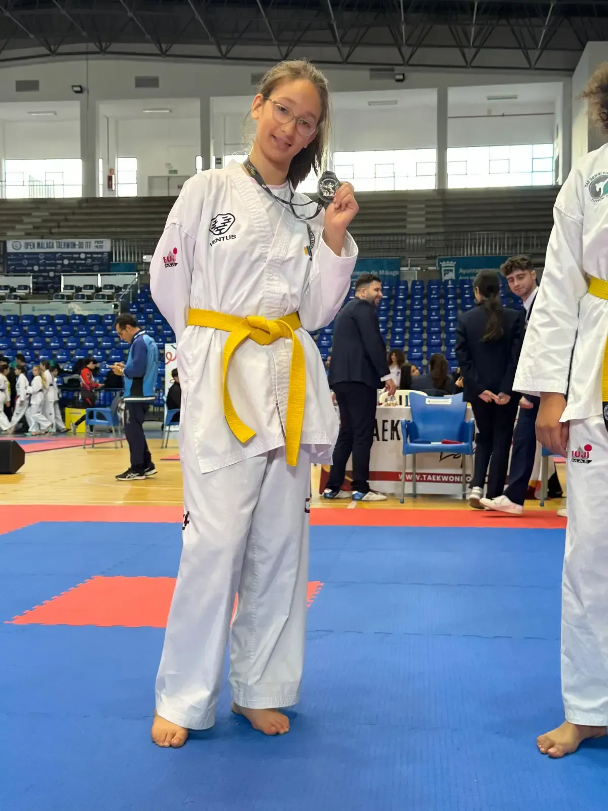 En un estadio, una persona sonriente, vestida con un uniforme blanco de taekwondo y un cinturón amarillo, se encuentra de pie