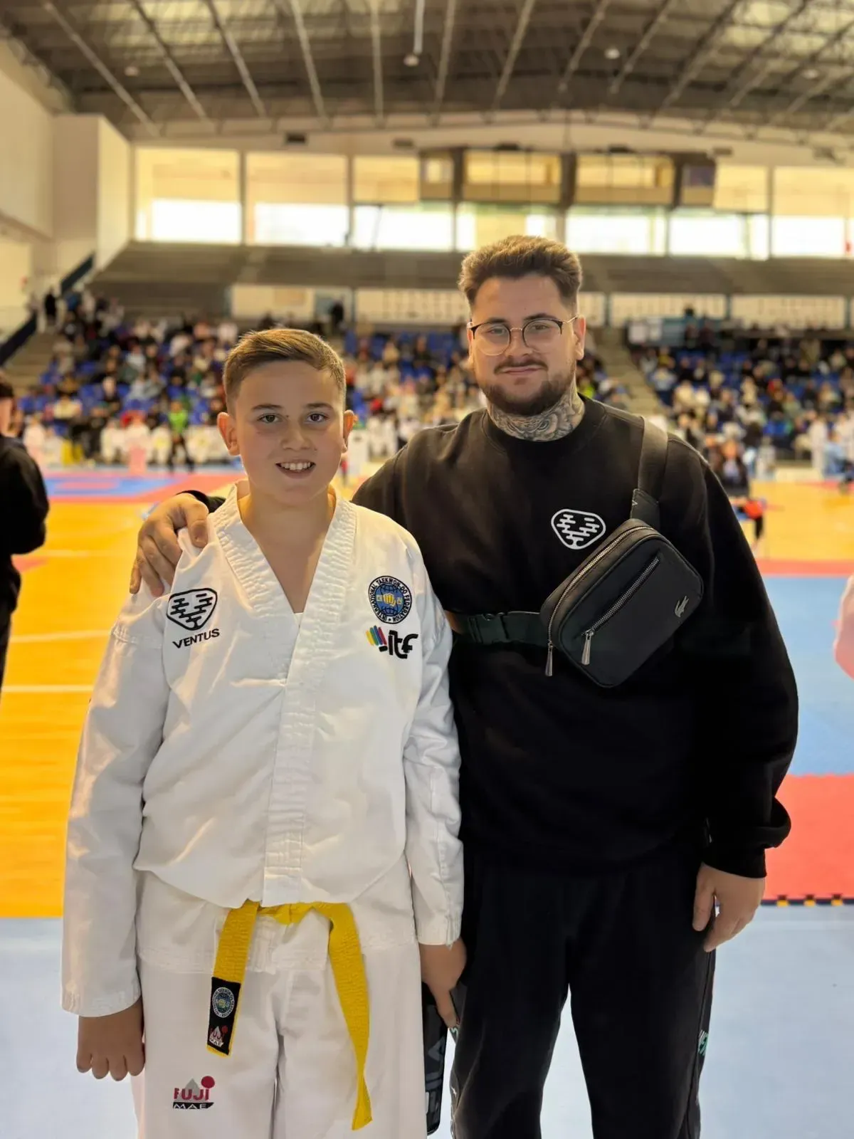 Un joven con un uniforme blanco de taekwondo y cinturón amarillo se encuentra de pie junto a un adulto