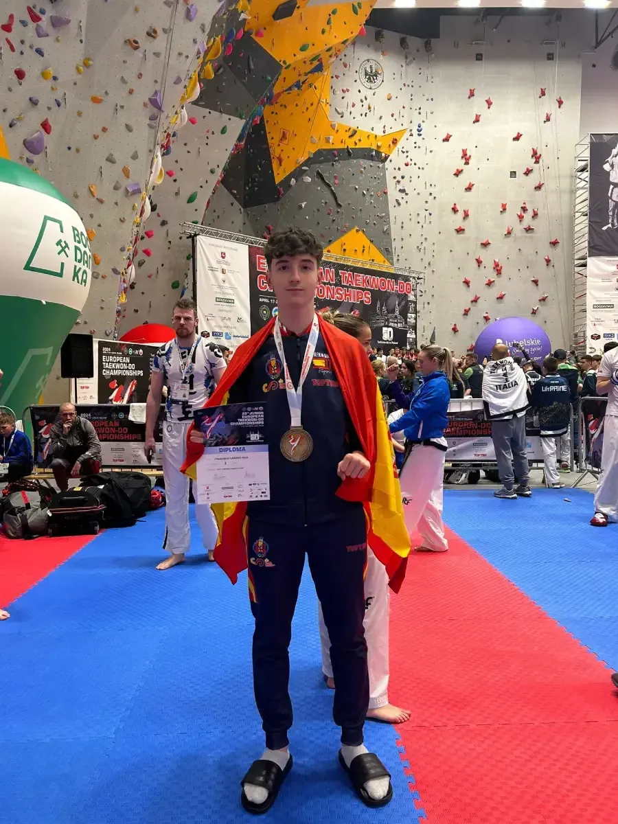 Una persona que lleva una medalla y una bandera española sobre los hombros sostiene un certificado en un rocódromo.