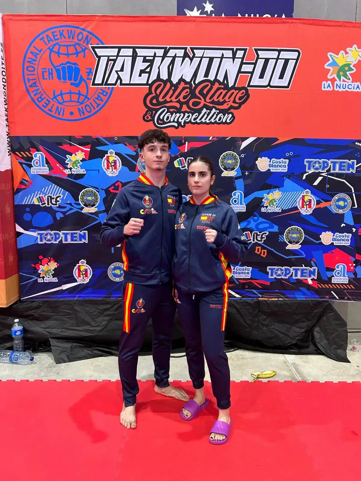 Dos atletas de taekwondo posan uno al lado del otro con chándales azul marino a juego, frente a un telón de fondo rojo y azu