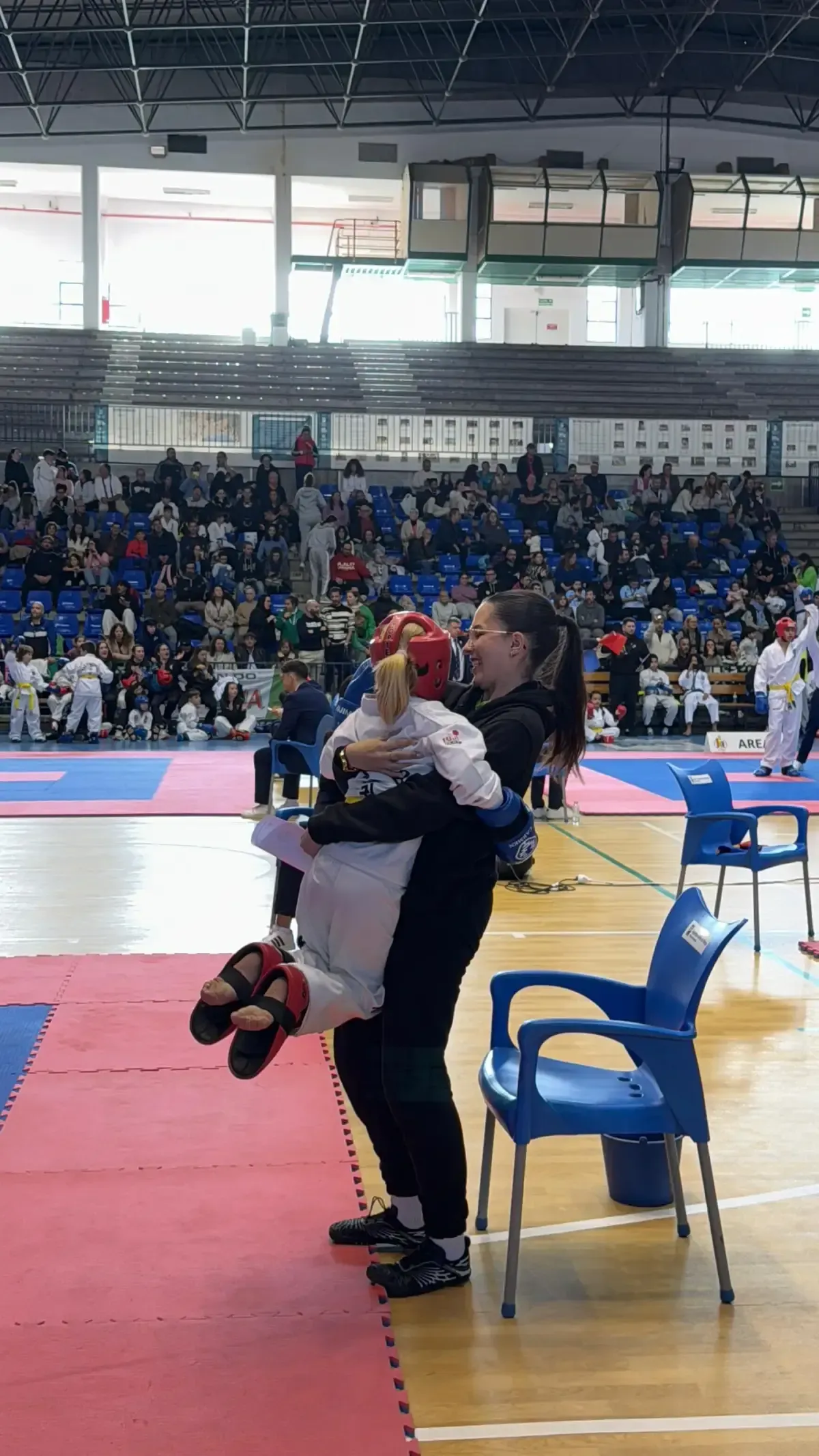 En un recinto cubierto, una persona vestida con uniforme negro sostiene a un niño con indumentaria de taekwondo