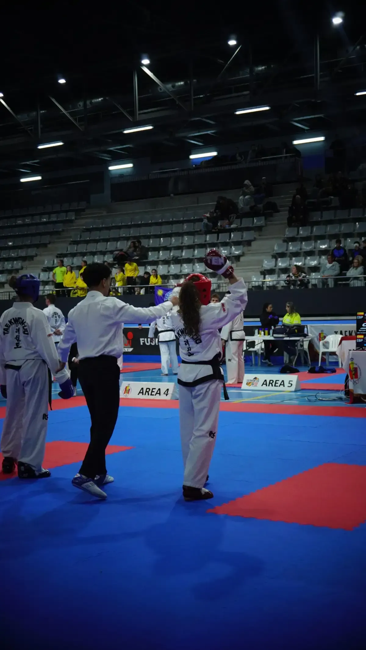 Un árbitro se sitúa entre dos competidores de taekwondo, equipados con indumentaria protectora, sobre una colchoneta