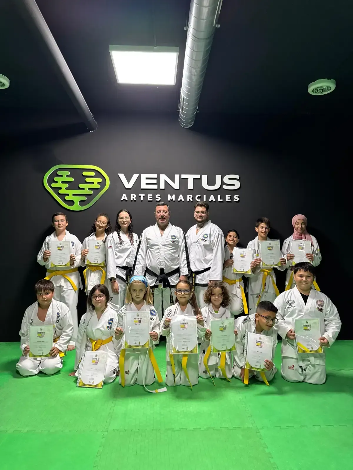 Un grupo de estudiantes e instructores con uniformes de artes marciales sosteniendo certificados en un estudio con una pared