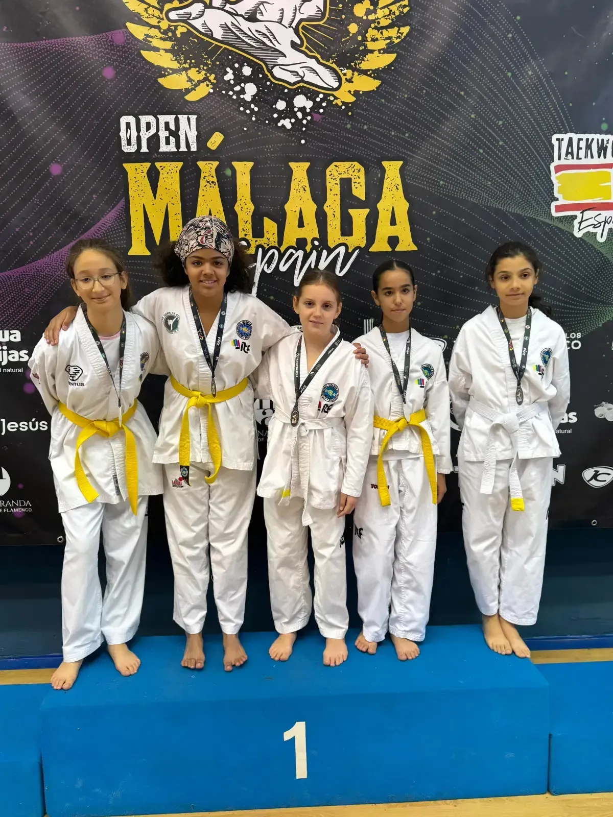 Cinco jóvenes competidores, vestidos con uniformes blancos de taekwondo y cinturones amarillos, se encuentran de pie