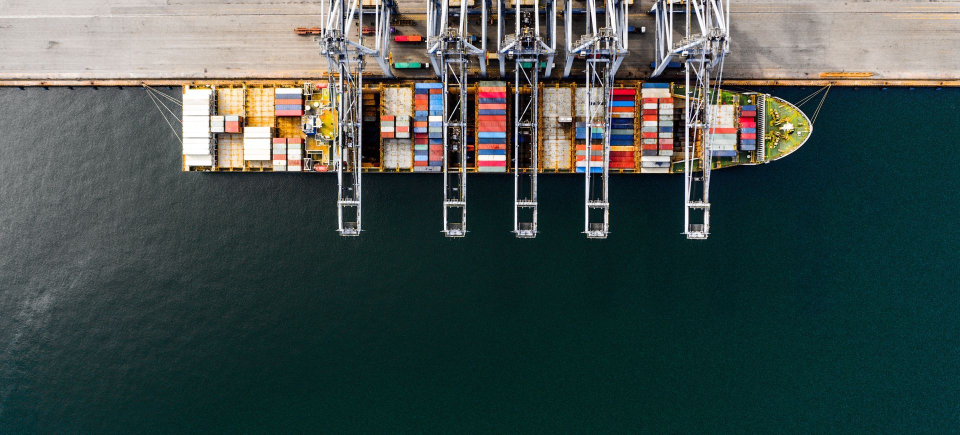 Vue aérienne d’un cargo amarré dans un port, entouré de grues et de conteneurs maritimes colorés.