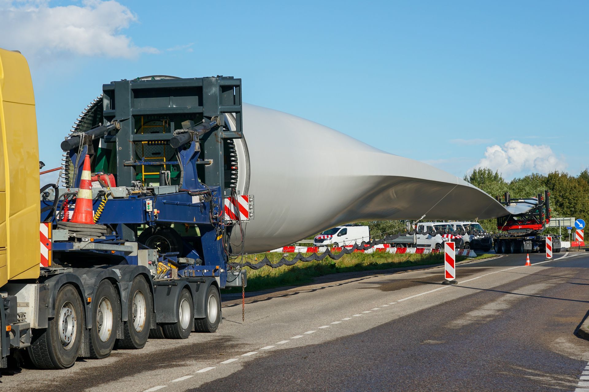 Une pale d’éolienne surdimensionnée transportée sur une remorque lourde le long d’une route balisée par des cônes de signalisation et accompagnée d’un véhicule d’escorte