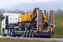 Un camion semi-remorque blanc transportant une grosse pelle jaune sur une remorque surbaissée, sur une route de campagne