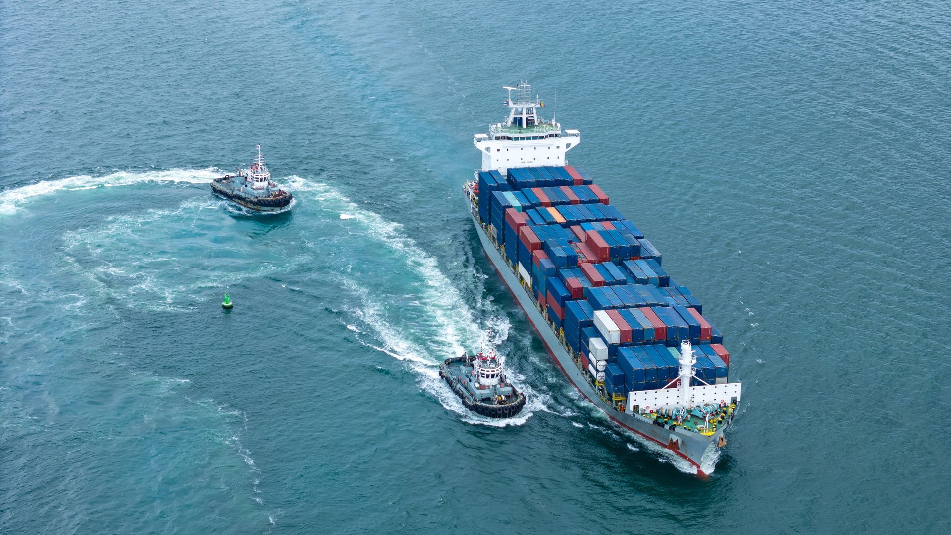 Vue aérienne d’un cargo remorquant un petit bateau et laissant un sillage blanc incurvé sur l’eau bleue.
