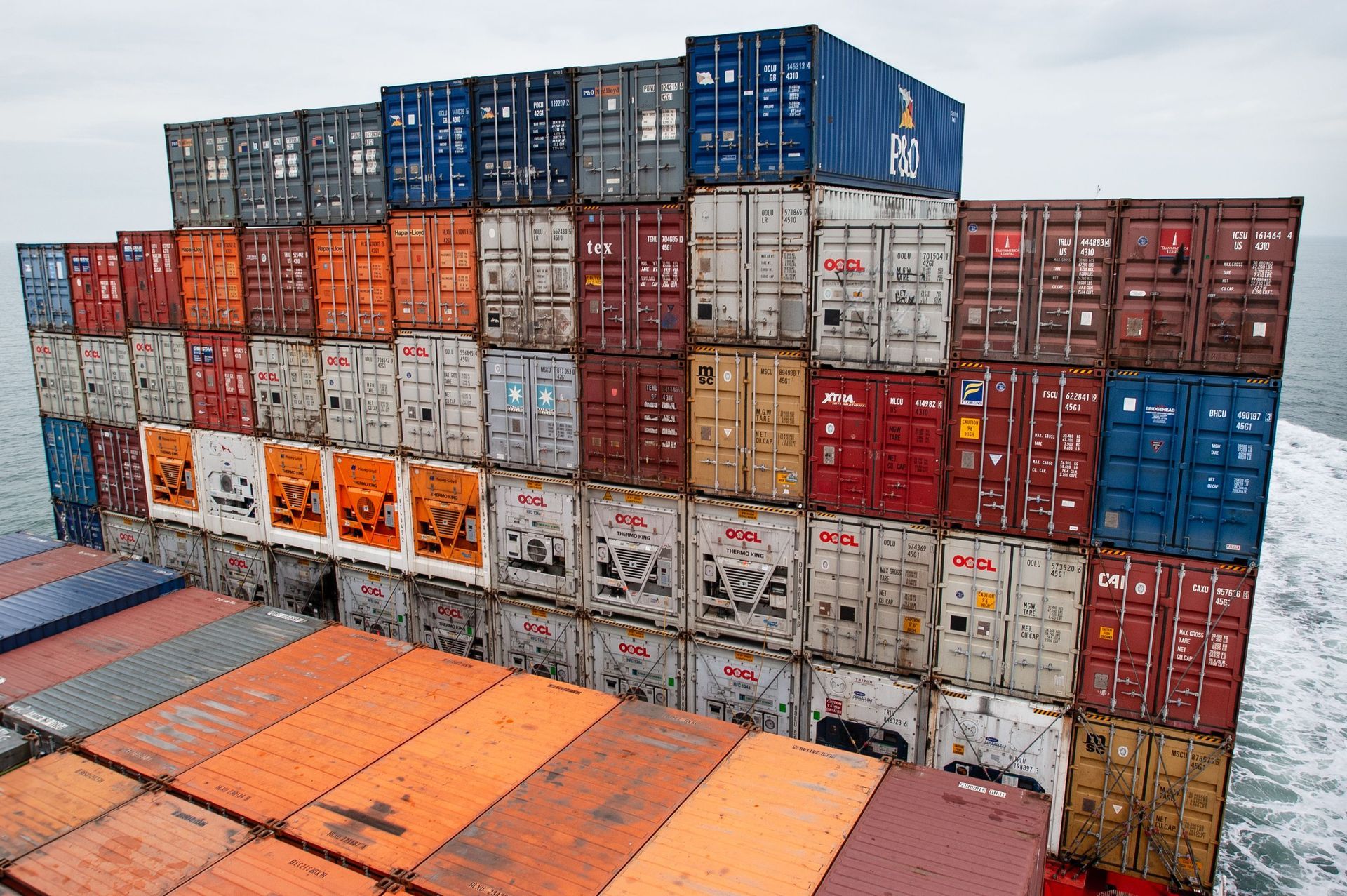Des piles de conteneurs maritimes bleus, rouges, blancs et orange dans un port, vues d’en haut.