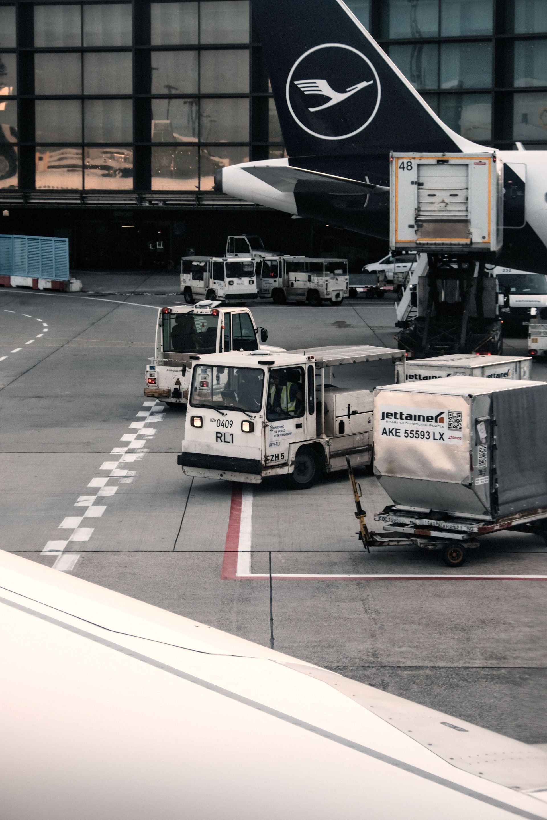 Des camions et du matériel de fret de Lufthansa sur le tarmac d’un aéroport, près de la queue d’un avion au terminal