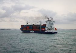 Un cargo navigue sur une eau calme sous un ciel nuageux, avec une coque rouge et bleue et une superstructure blanche.