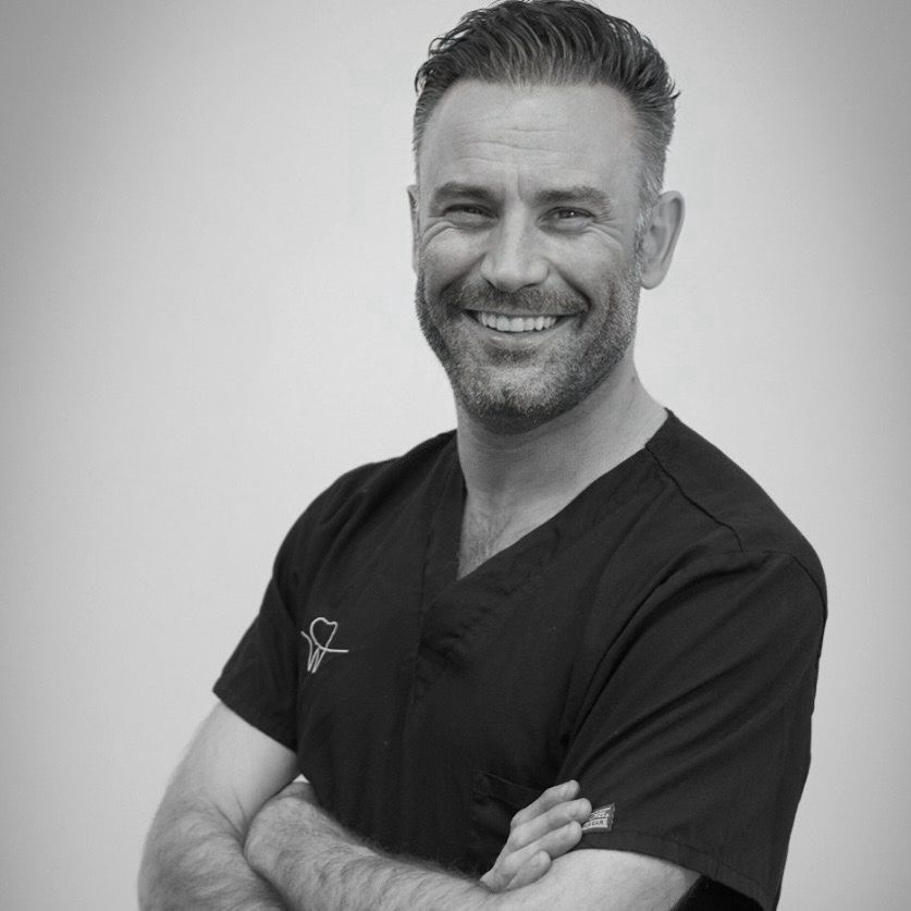 Hombre sonriente con uniforme negro y brazos cruzados sobre un fondo blanco.