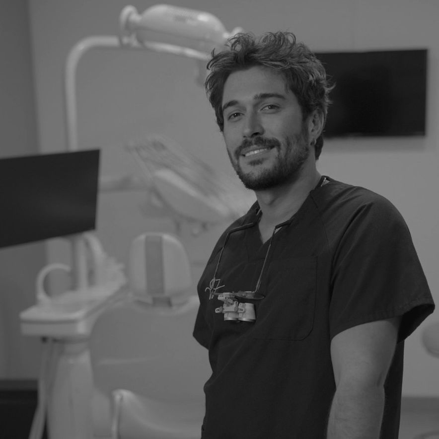 Dentista sonriendo en un consultorio dental; equipo médico en el fondo.