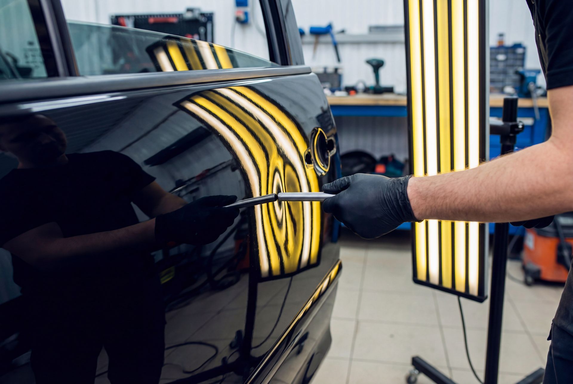 Un ouvrier ganté polit une portière de voiture noire dans un garage, sous des reflets de lumière jaune.