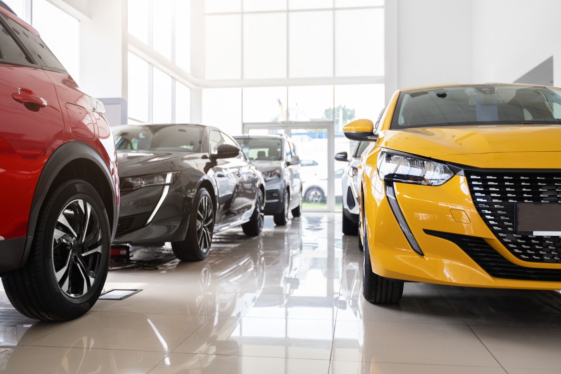 Salle d'exposition automobile lumineuse avec une berline sport jaune, une voiture noire et un SUV rouge sur un sol brillant.