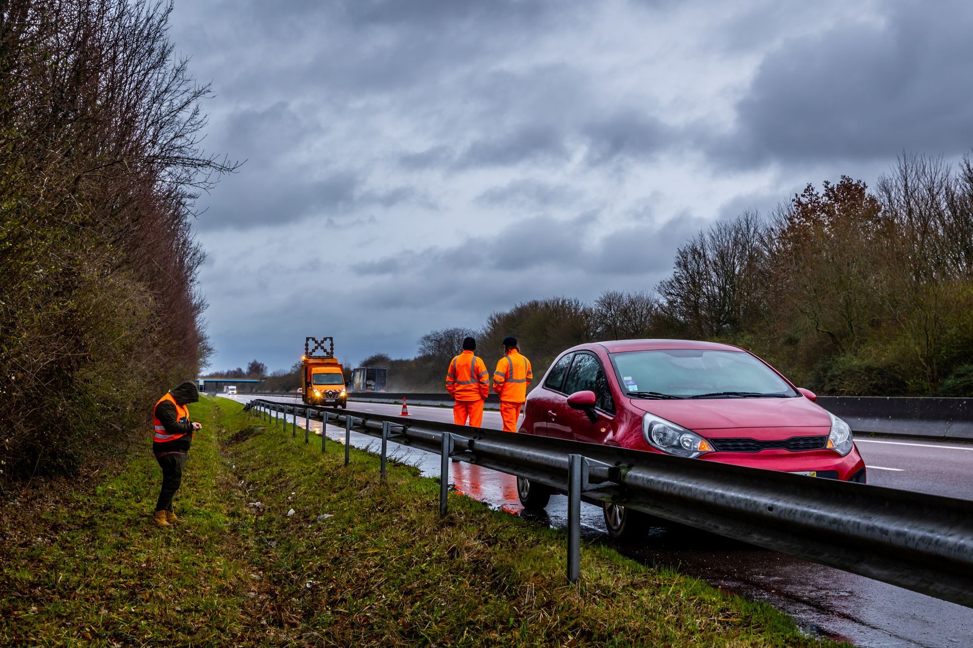 Voiture rouge contre la glissière de sécurité, secouristes en uniforme orange près de la chaussée sous un ciel nuageux.