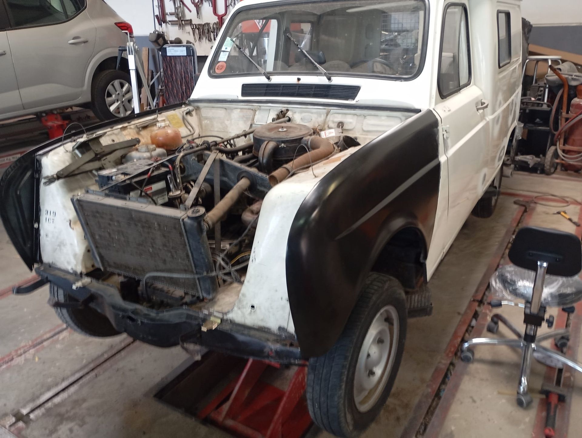 Une vieille voiture blanche, dont les panneaux de carrosserie avant ont été retirés, se trouve dans un garage, laissant apparaître le moteur et le châssis.