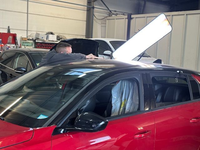 Voiture rouge dans un garage, capot ouvert, un mécanicien travaille dessous.