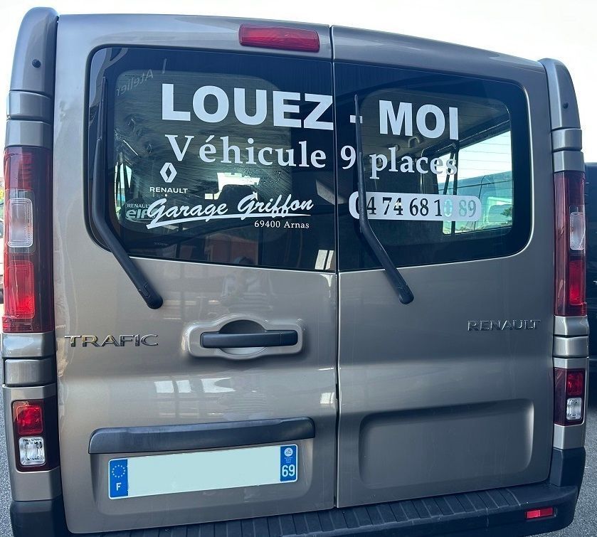 Fourgonnette argentée avec panneau de location et vitres arrière recouvertes de texte.