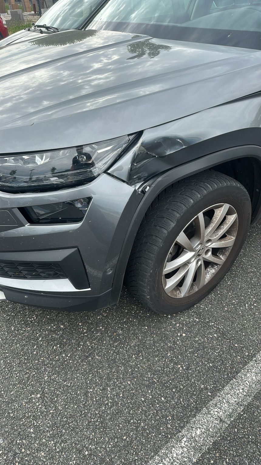 SUV gris stationné sur un parking, vue avant gauche avec des dommages visibles aux roues et au capot, et chaussée mouillée.
