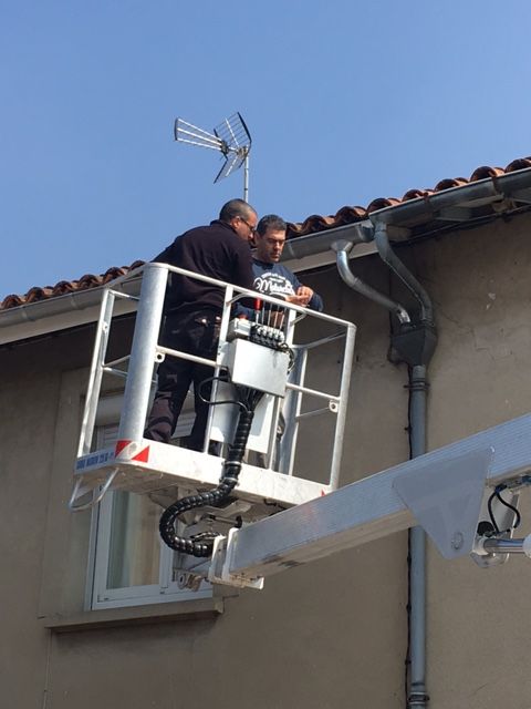 Deux ouvriers, installés dans une nacelle blanche, inspectent une gouttière et une antenne sur le toit d'une maison.