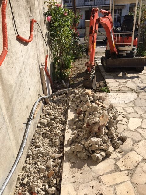 Une pelleteuse dégage des gravats le long d'une allée en pierre, près d'un mur et d'un jardin.