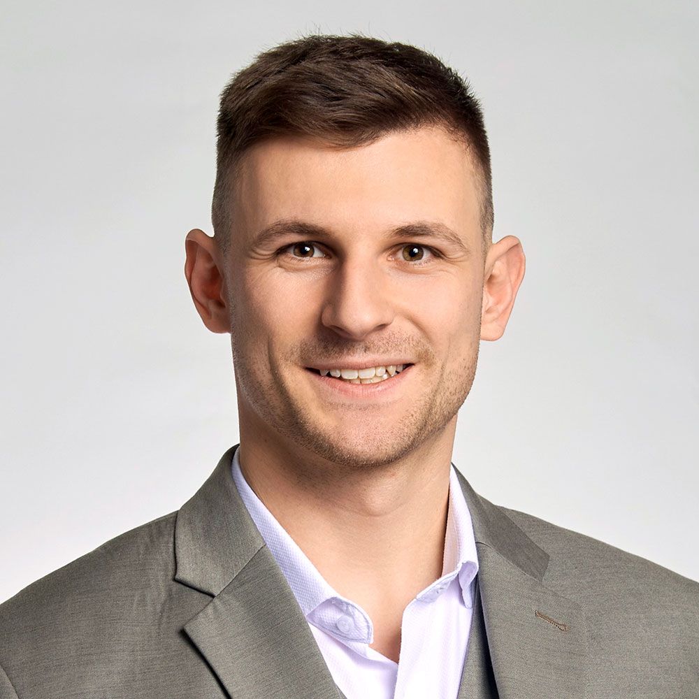 Ein Mann in Anzug und Krawatte steht vor einem grauen Hintergrund.