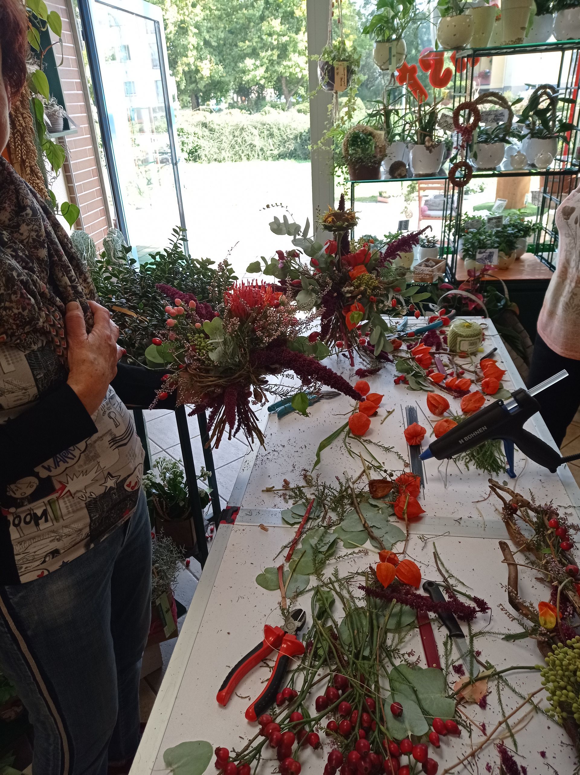 Menschen arrangieren Blumen an einem Floristentisch. Rote und orangefarbene Blüten, Grünpflanzen und Werkzeuge sind zu sehen.