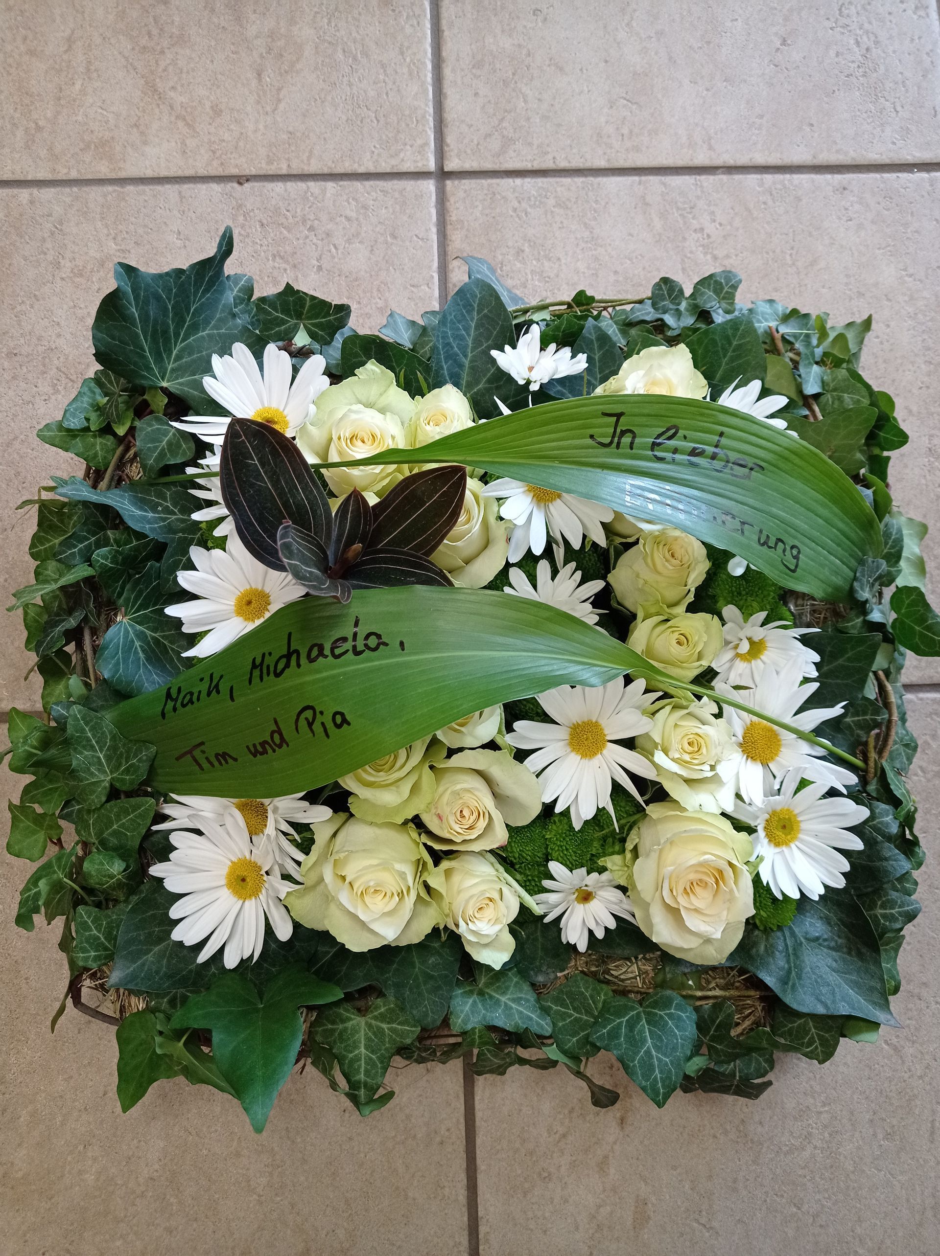 Trauerblumenarrangement mit weißen Rosen und Gänseblümchen, grünem Efeu, einem Schmetterling und zwei Inschriften auf Blättern.