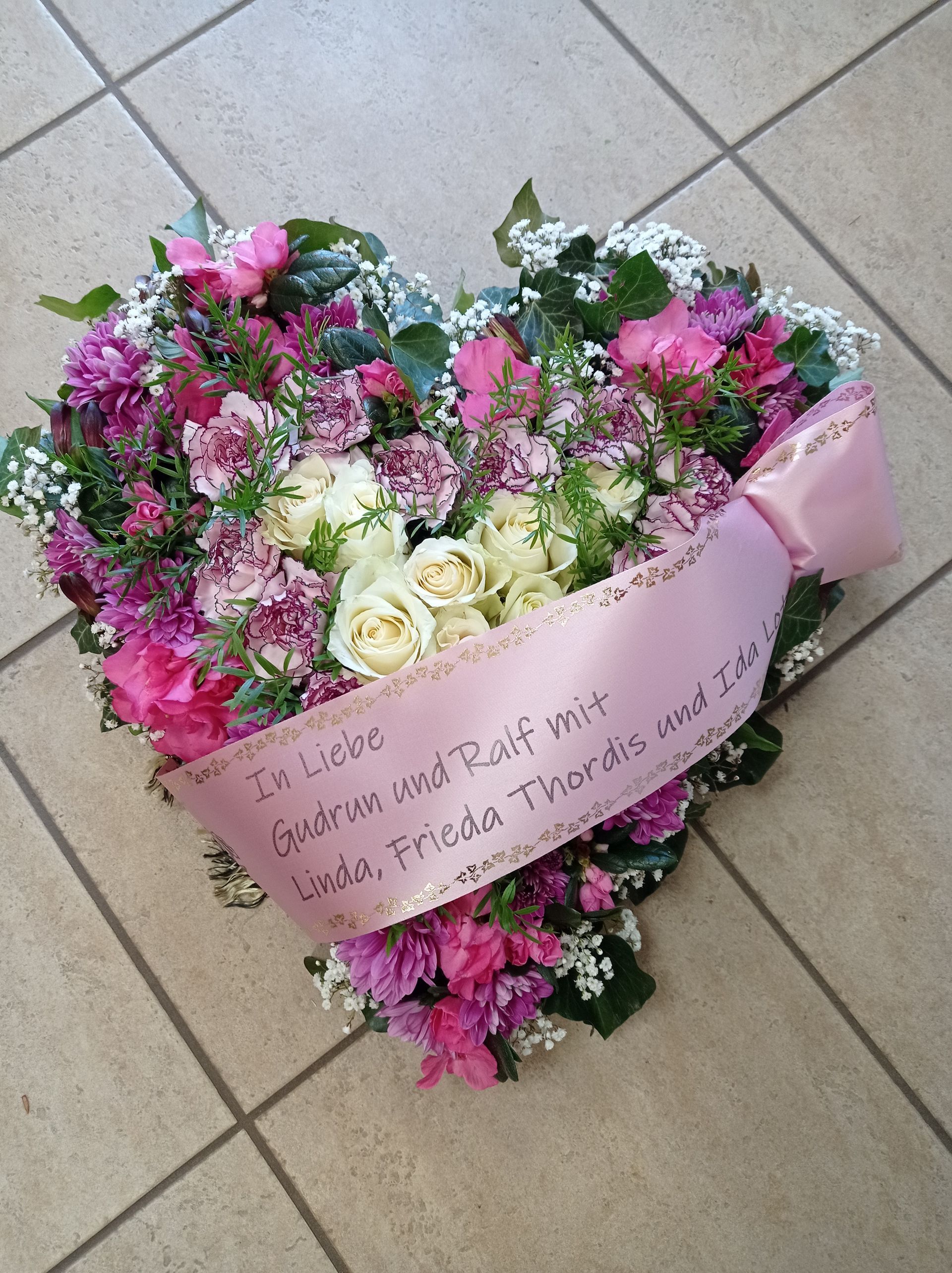 Herzförmiges Blumenarrangement mit rosa Schleife. Text auf der Schleife: „In Liebe…“, Blumen: rosa, weiß, grün, auf gefliestem Boden.