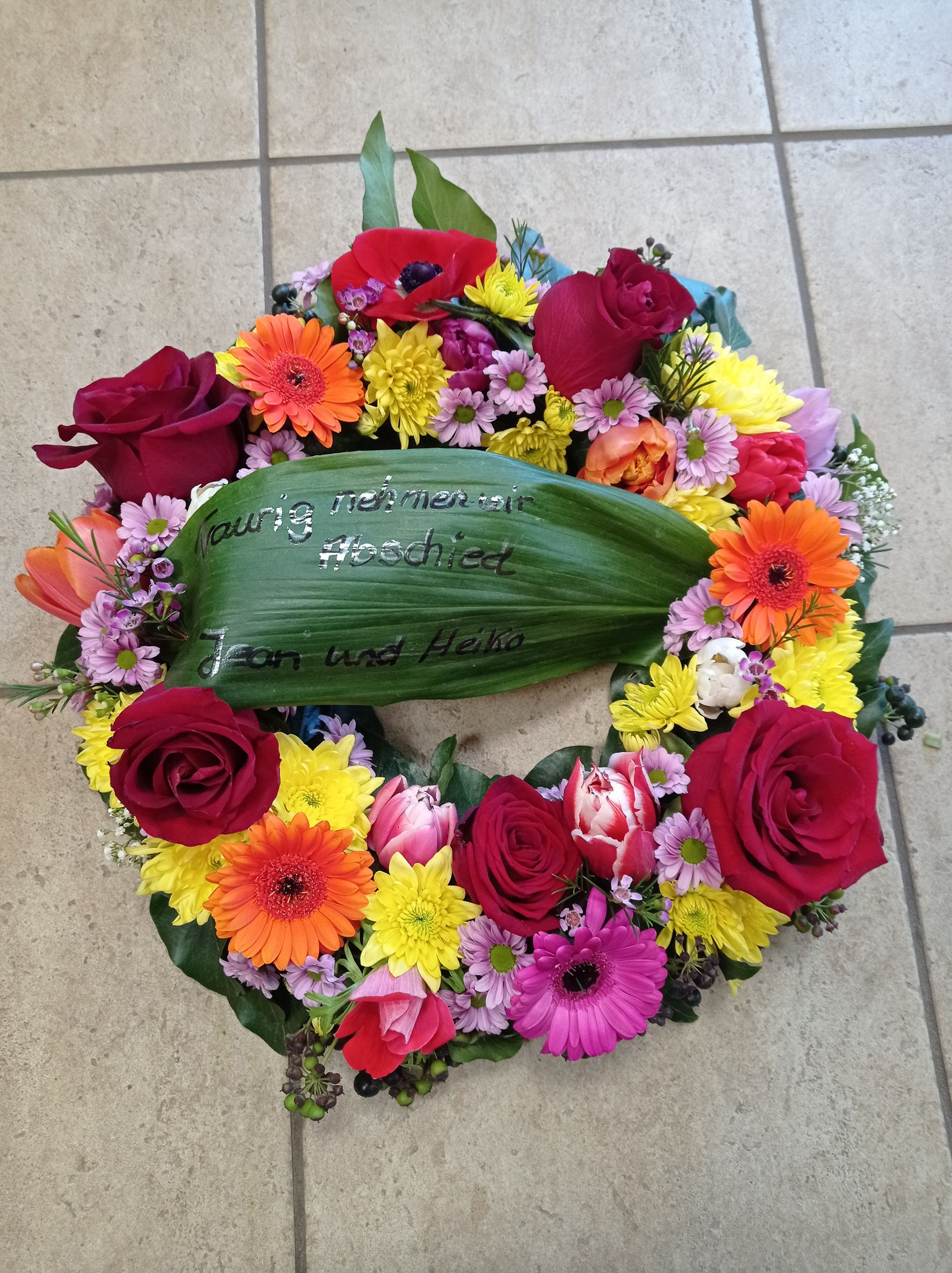 Bunter Blumenkranz mit roten Rosen, orangefarbenen Gerbera-Gänseblümchen und einem grünen Blatt mit Aufschrift.