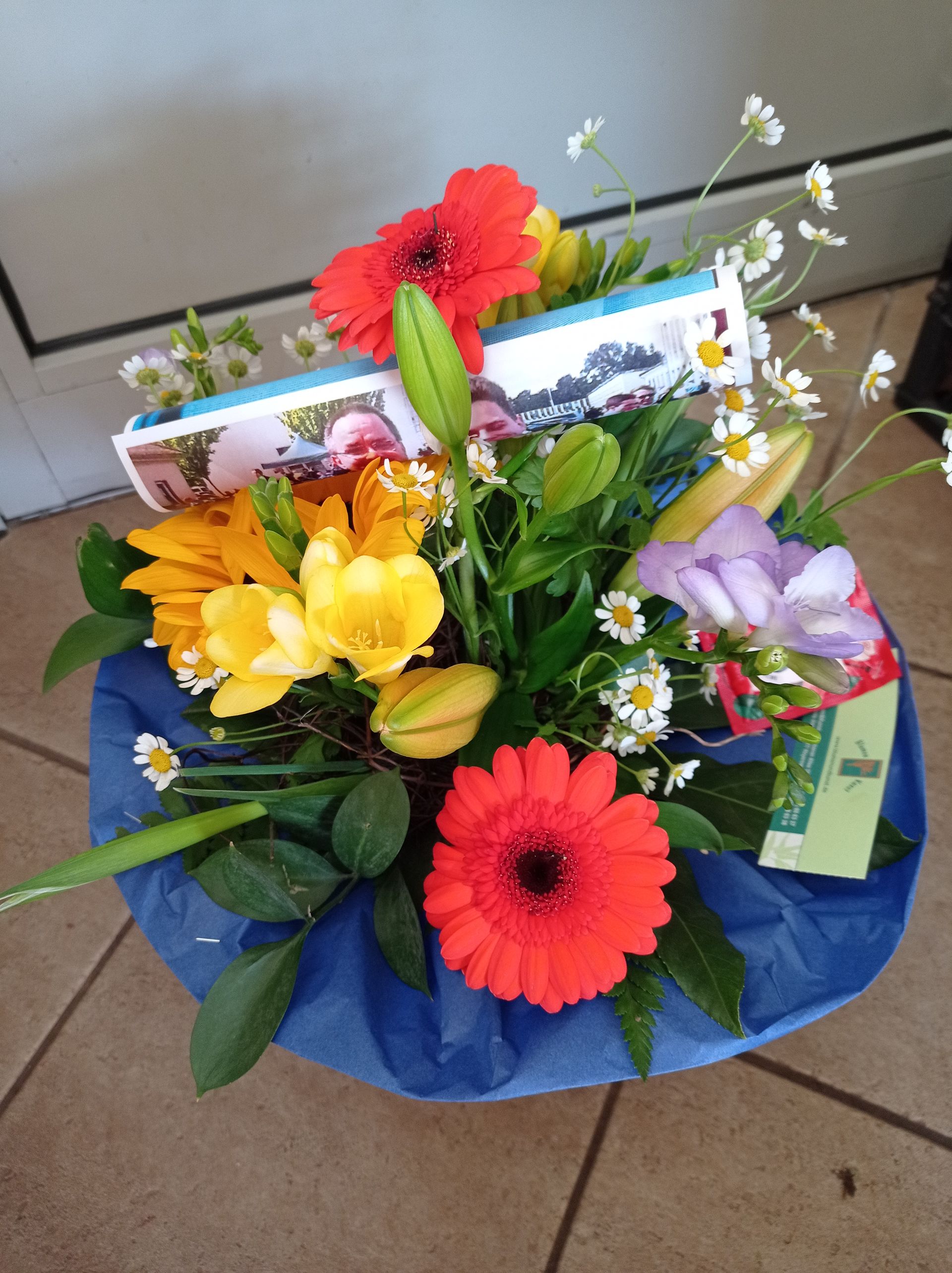 Blumenstrauß mit orangefarbenen Gerbera-Gänseblümchen, gelben Freesien und einer in blaues Papier eingewickelten Geschenkkarte.