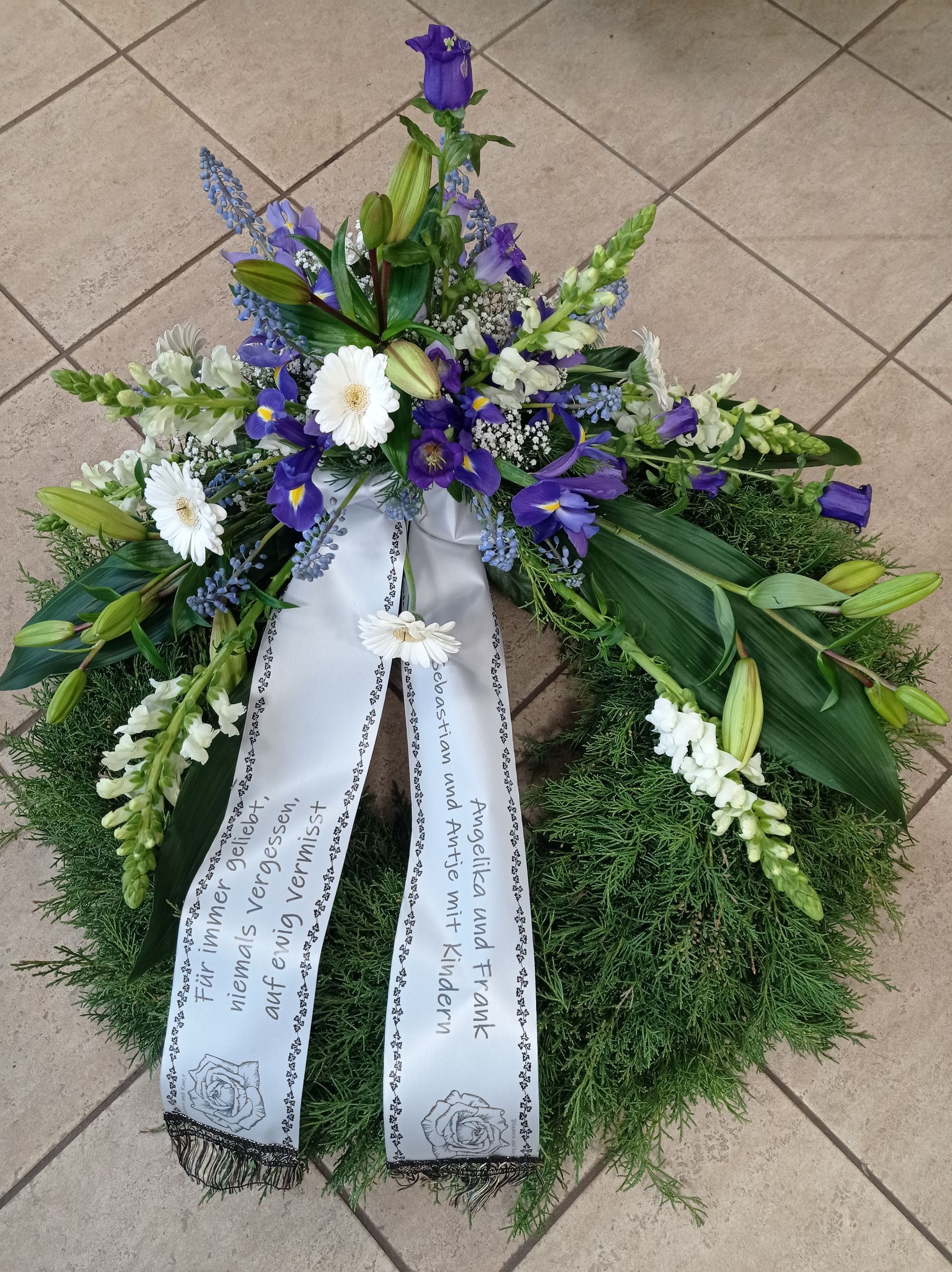 Kranz aus grünem Laub und blauen und weißen Blumen, mit einem weißen Band, wahrscheinlich für ein Denkmal.