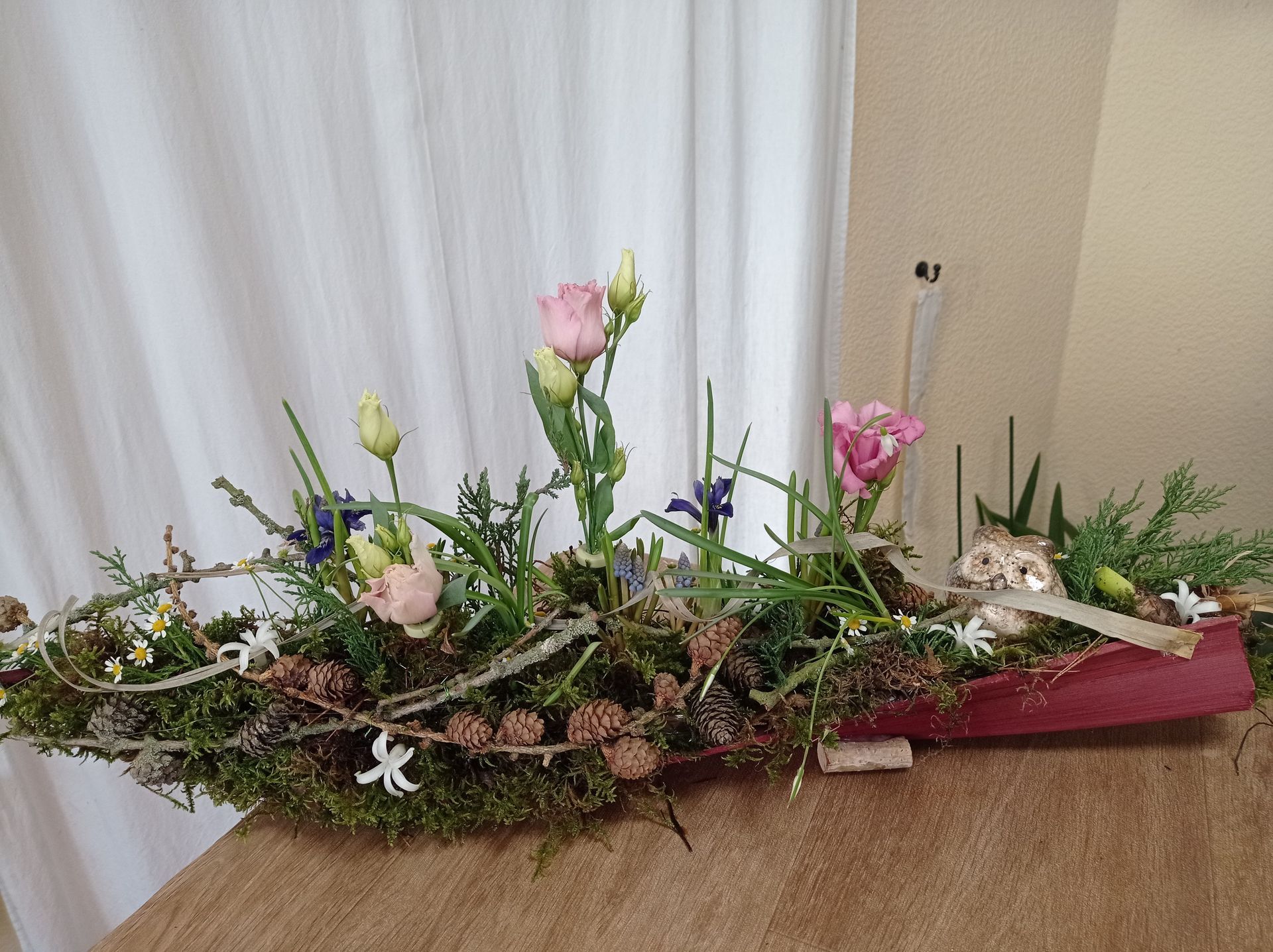 Blumendekoration mit rosa Blumen, Grünpflanzen und kleinen Zapfen in einem langen, burgunderfarbenen Behälter.