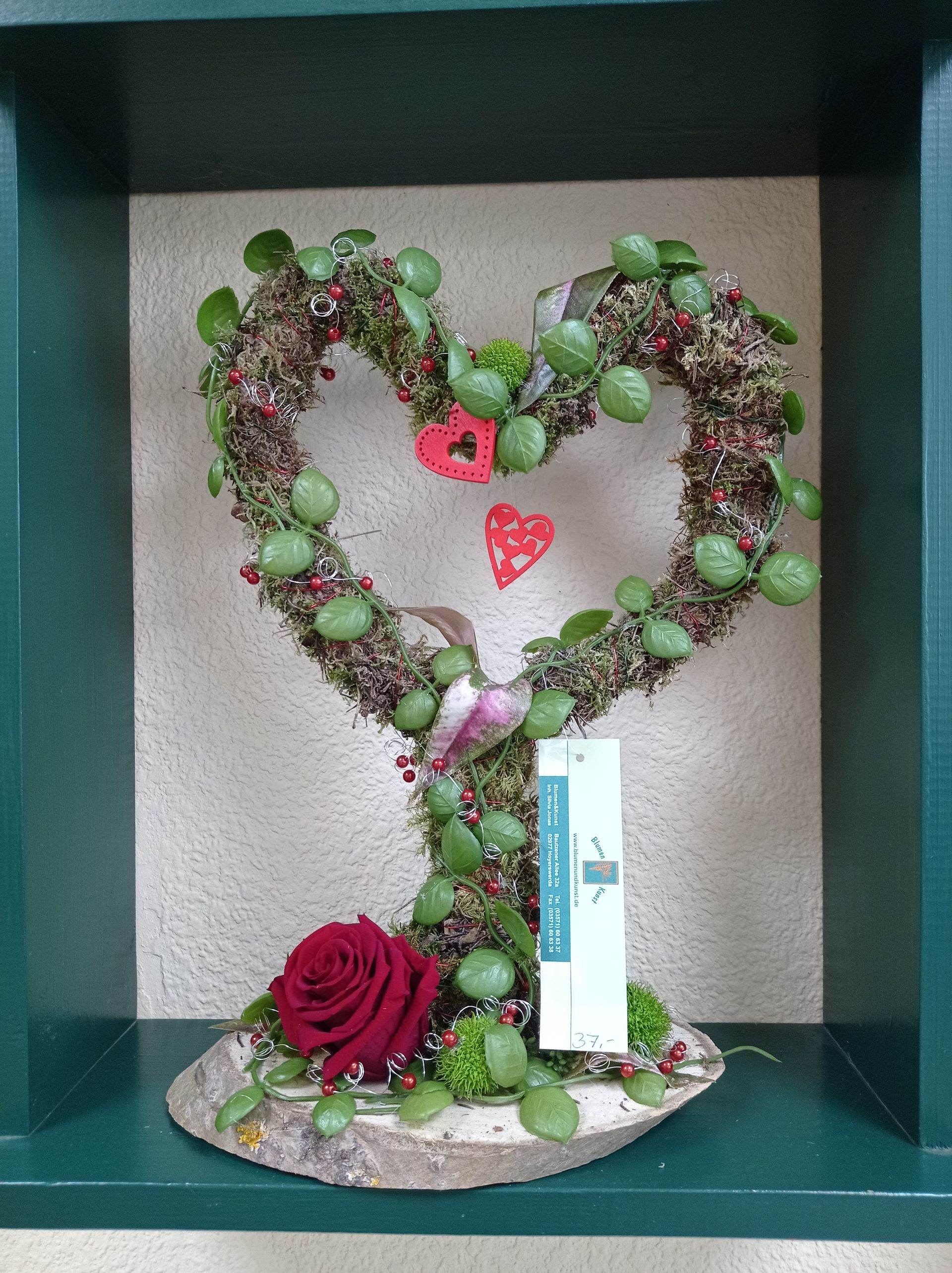 Herzförmiges Blumenarrangement mit einer roten Rose, Moos und Grünpflanzen. Präsentiert in einer grün gerahmten Nische.