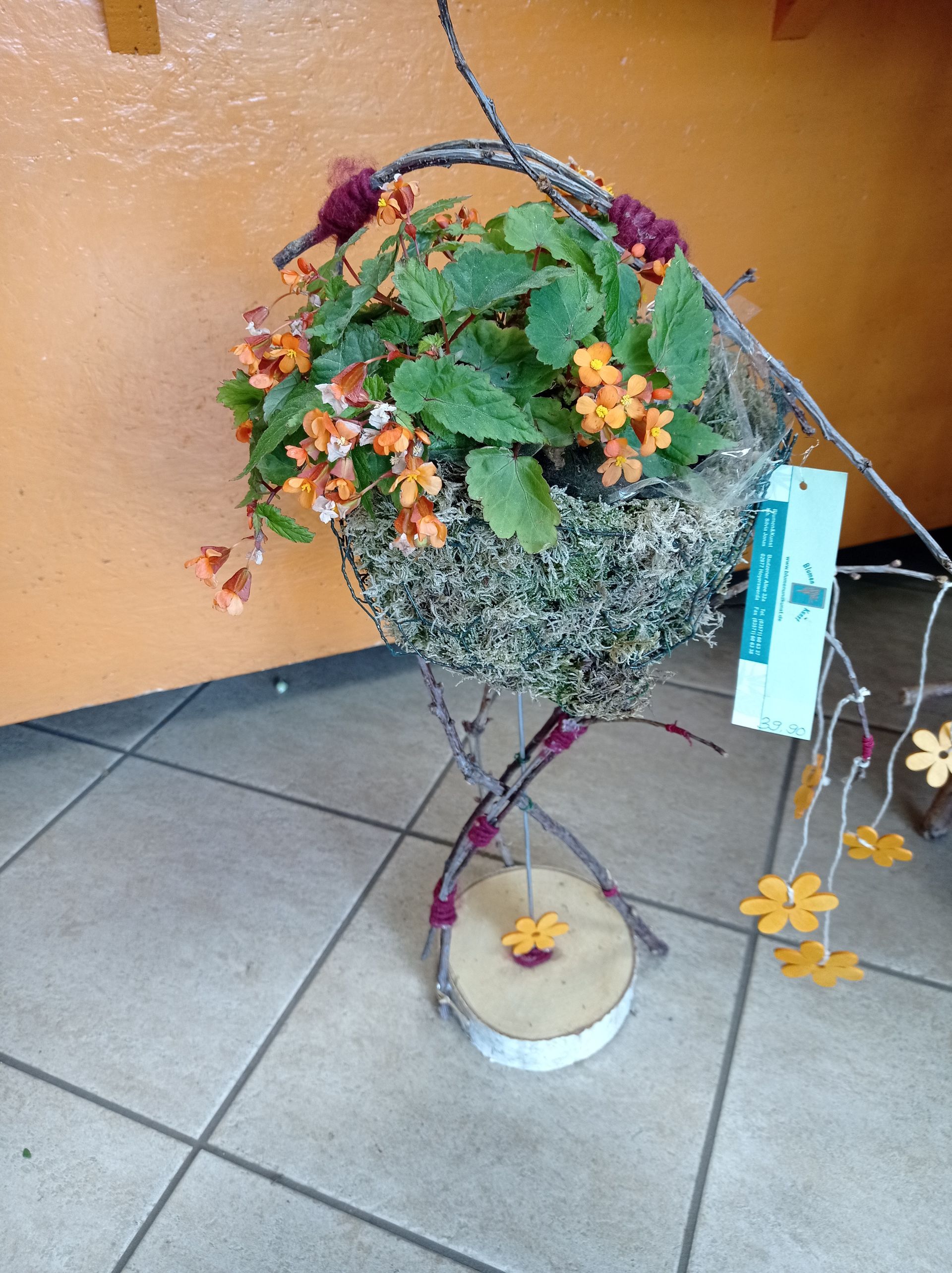 Arrangement: Orangefarbene Blüten und grüne Blätter in einem nestartigen Pflanzgefäß, getragen von Ästen, mit Dekor und Holzsockel.