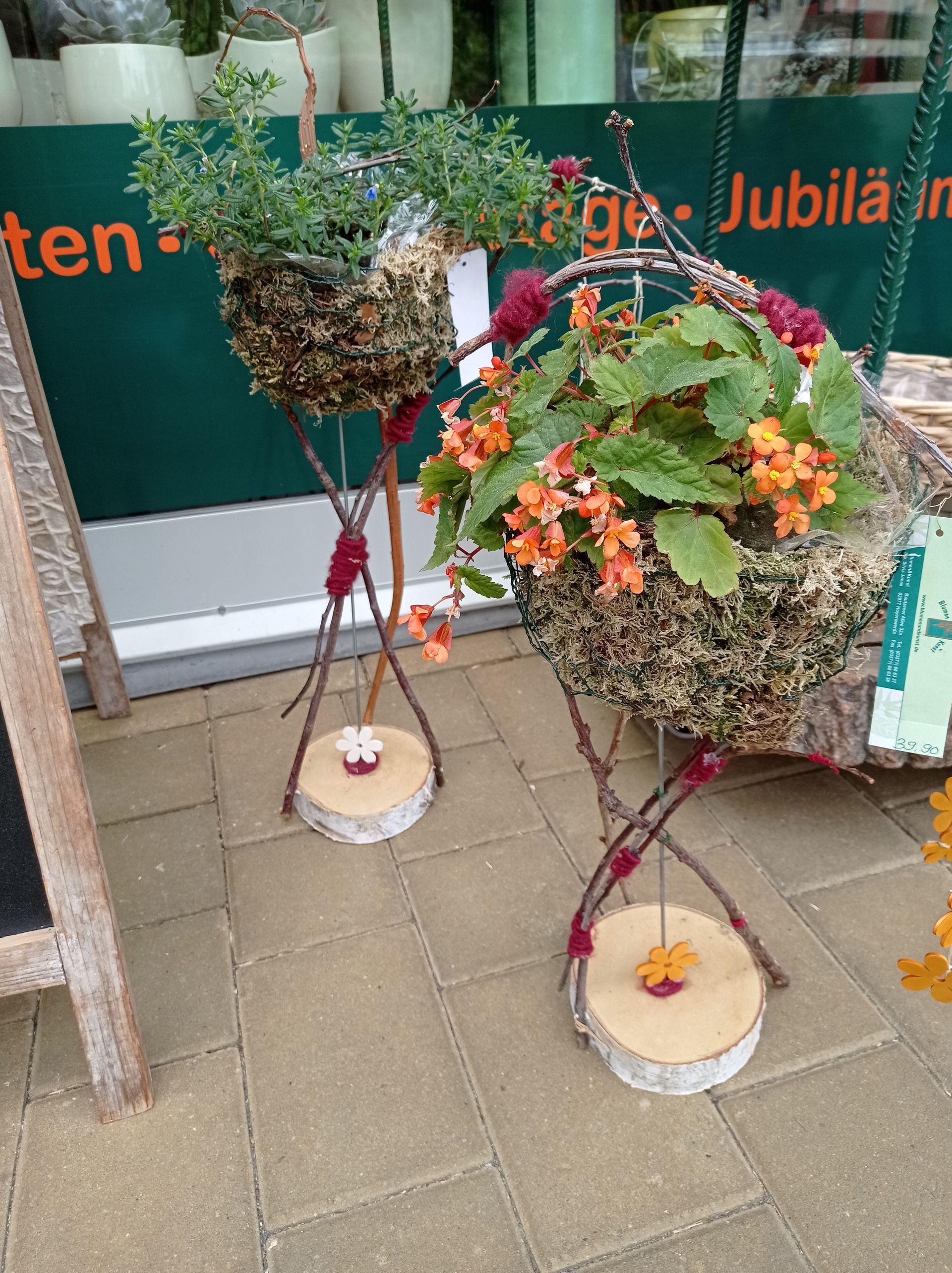 Zwei hängende Blumenkörbe mit bunten Blüten, die auf Holzsockeln stehen, vor einem Geschäft.