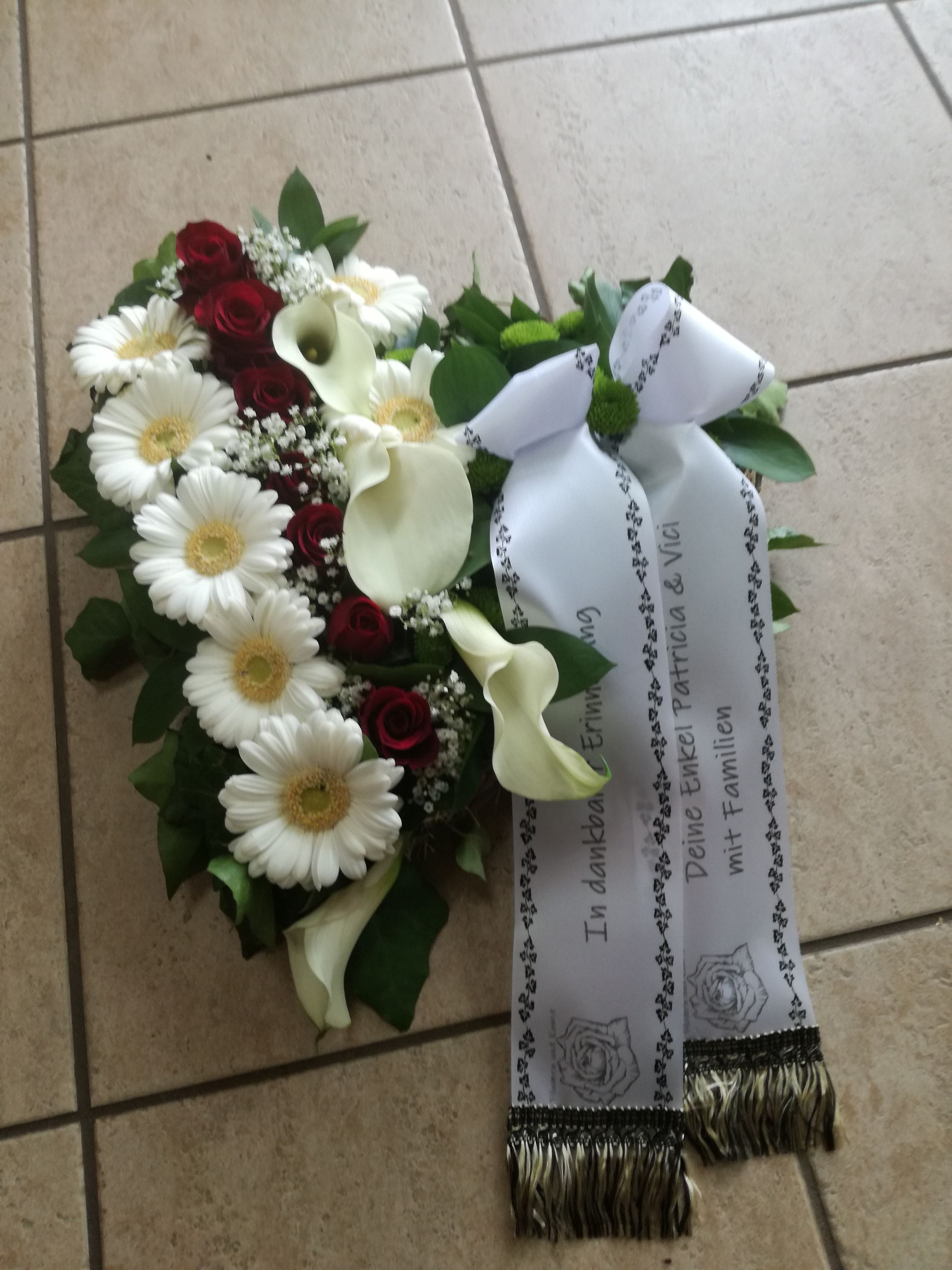 Herzförmiges Blumenarrangement: weiße Gänseblümchen, Calla-Lilien, rote Rosen und Band mit Text auf gefliestem Boden.