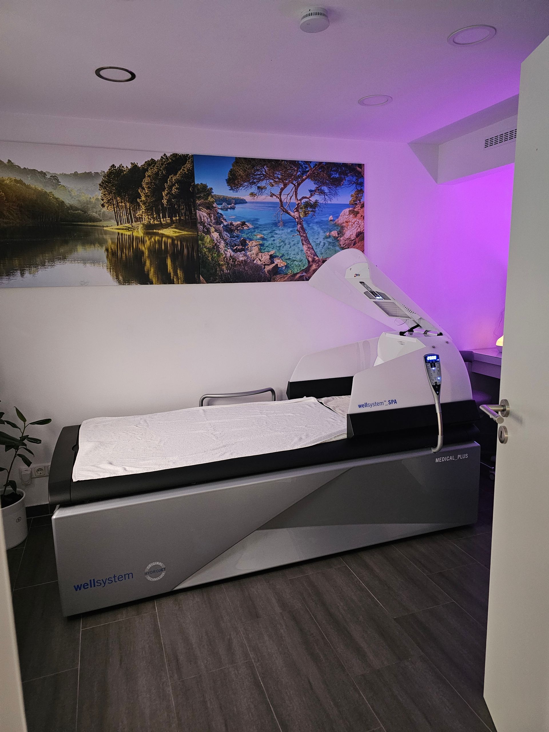 Behandlungsraum mit Krankenliege, Untersuchungsarm, Landschaftsbildern an der Wand und violetter Akzentbeleuchtung.