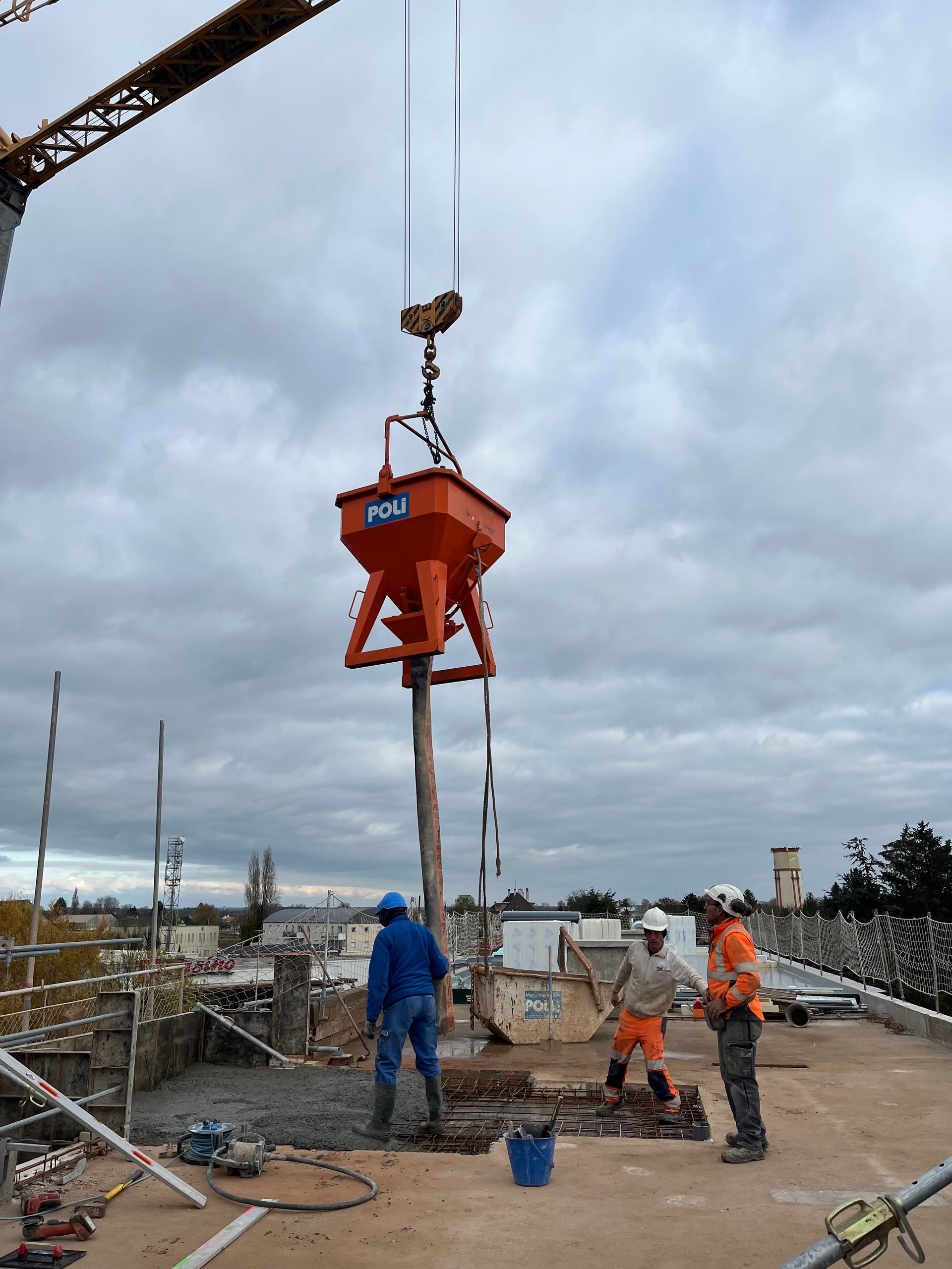 Grue de construction soulevant un conteneur rouge sur un chantier. Des structures en métal et des matériaux de construction sont visibles.