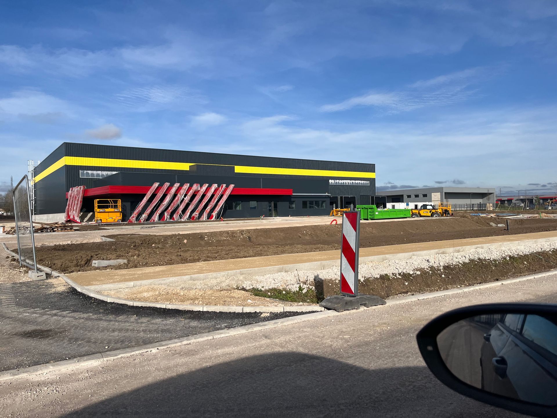 Bâtiment industriel moderne avec des structures en métal et un parking en face. Le ciel est clair et ensoleillé.