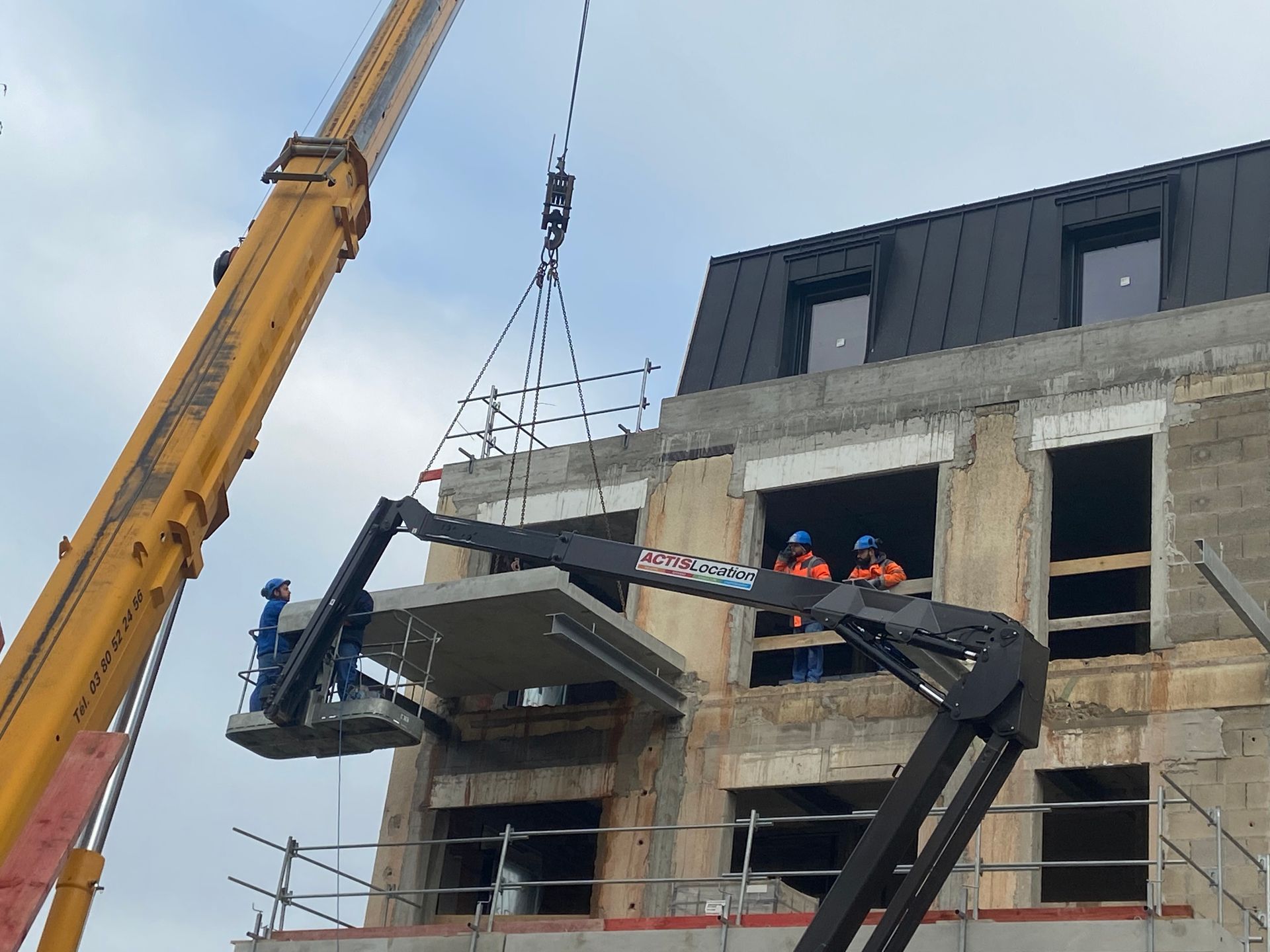 Grue de construction soulevant des matériaux sur un chantier. Des ouvriers sont visibles en haut de la grue, et le bâtiment en arrière-plan est partiellement construit.