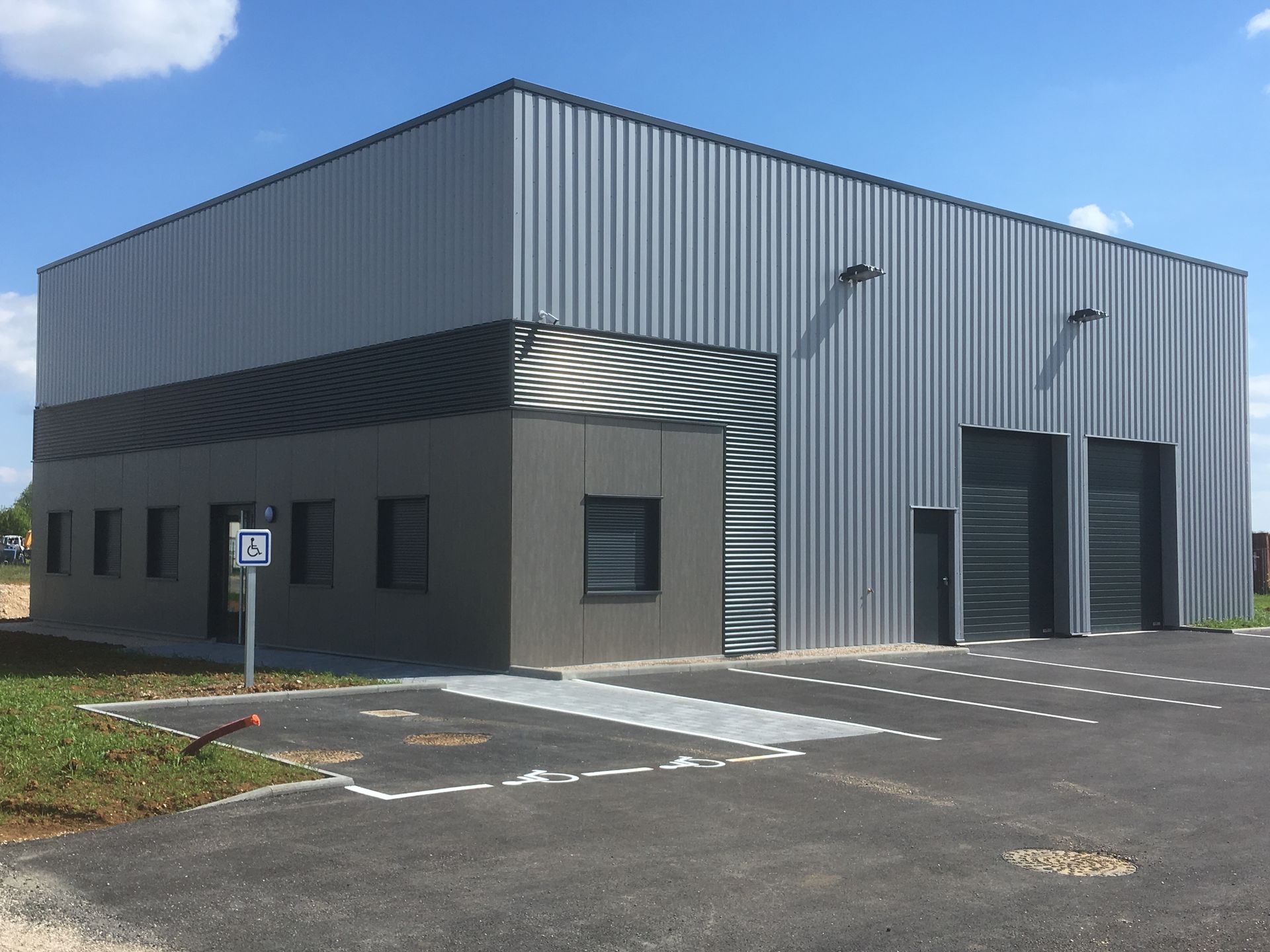 Bâtiment industriel moderne avec une façade en métal et des fenêtres. Le bâtiment est entouré d'un parking vide et le ciel est clair et ensoleillé.
