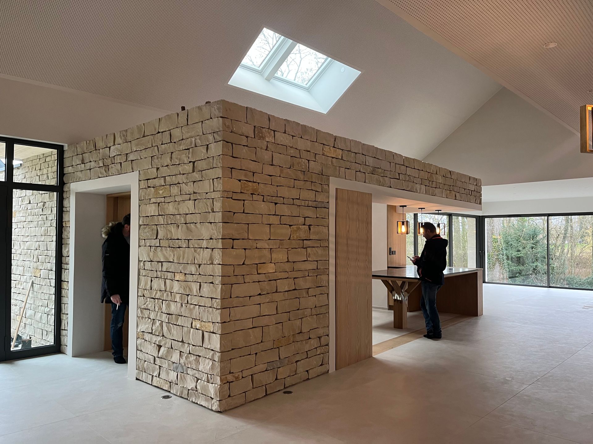 Intérieur d'un bâtiment moderne avec un espace d'accueil en pierre apparente et des baies vitrés derrière.