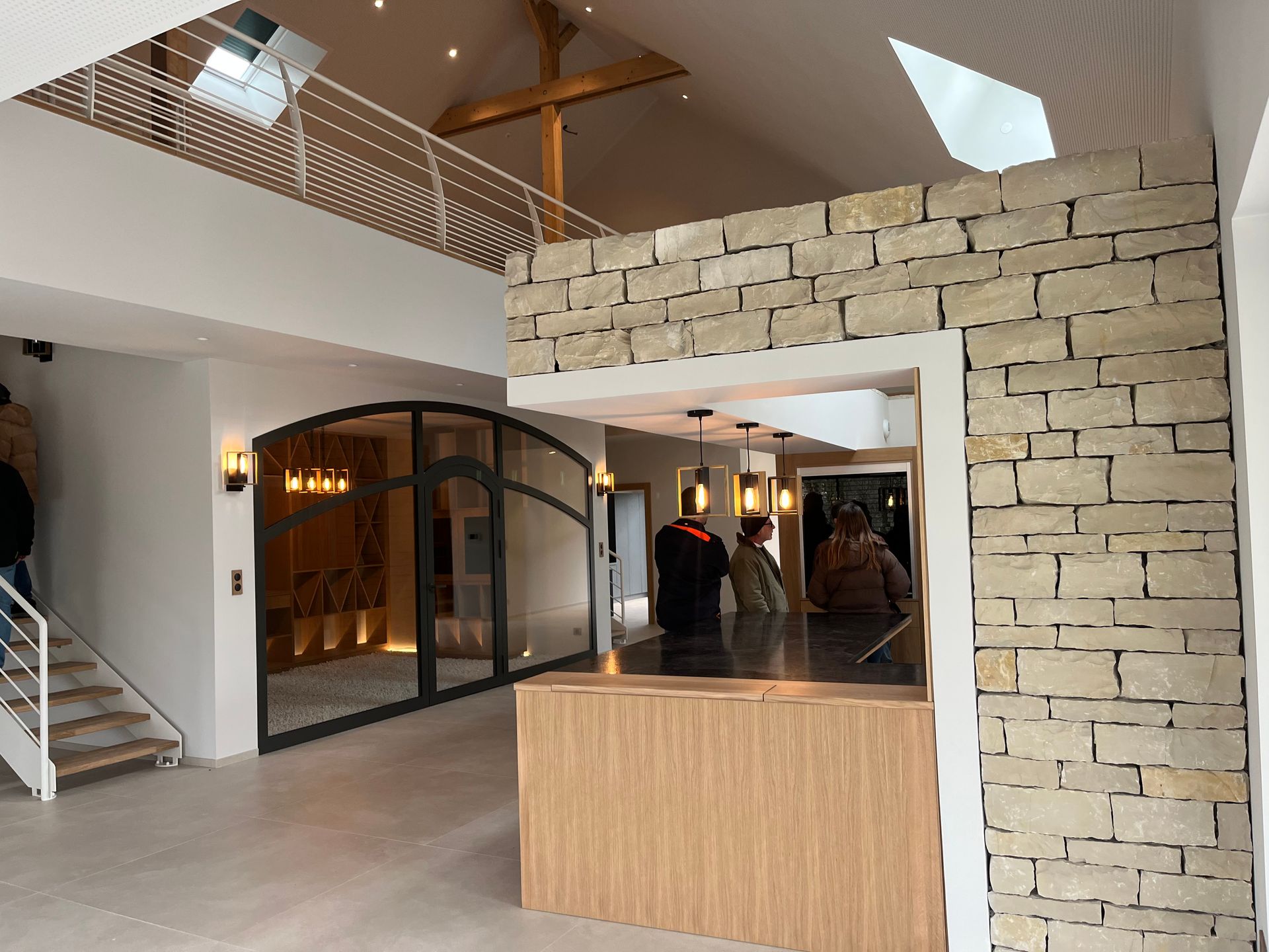 Intérieur d'un bâtiment moderne avec un espace d'accueil en pierre apparente et une baie vitré en métal noir. La pièce est bien éclairée et un espace de vie.