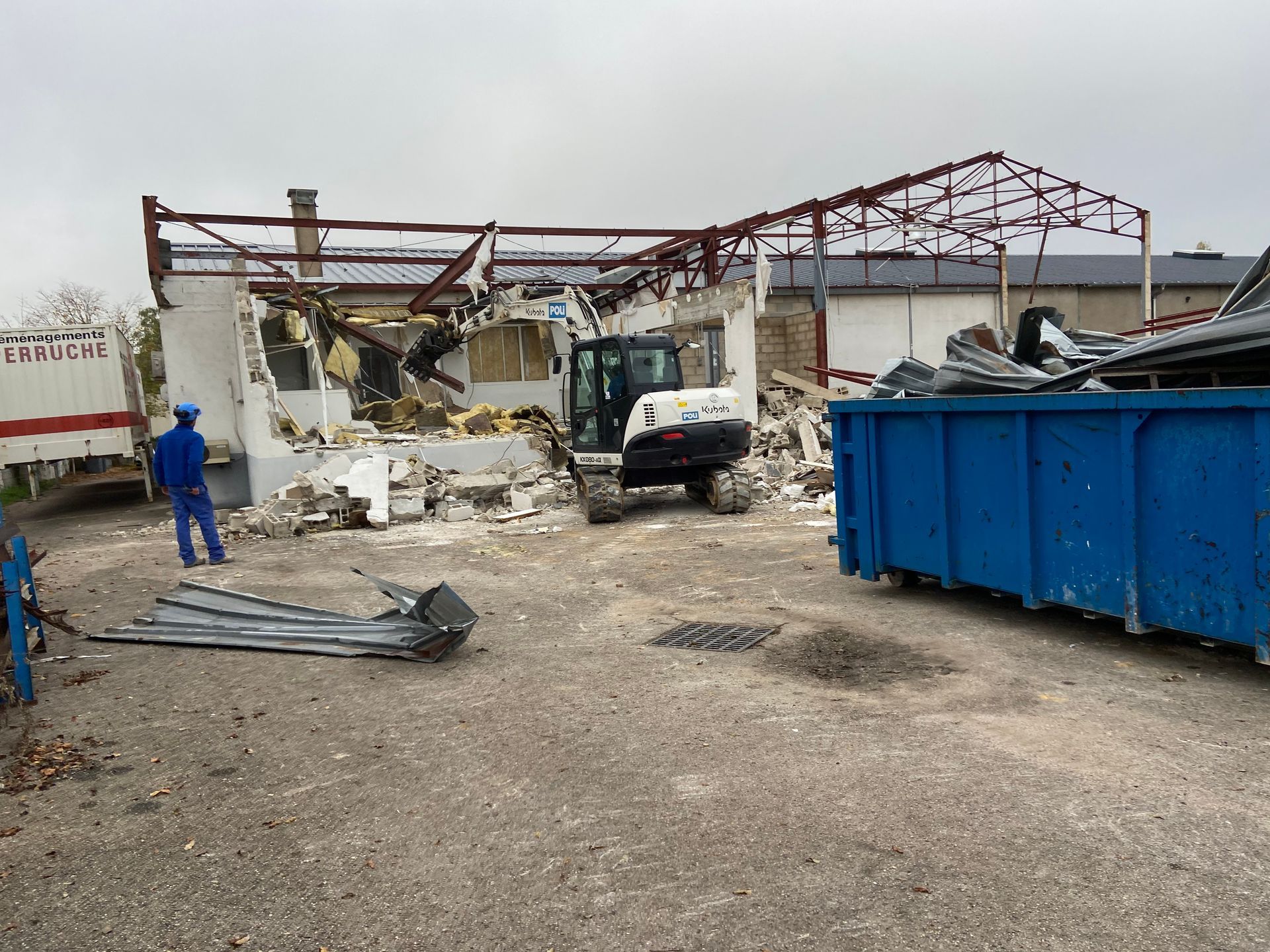 Chantier de démolition avec des débris de construction et un engin de chantier. Des ouvriers sont visibles, et un conteneur bleu est présent pour l'évacuation des débris.