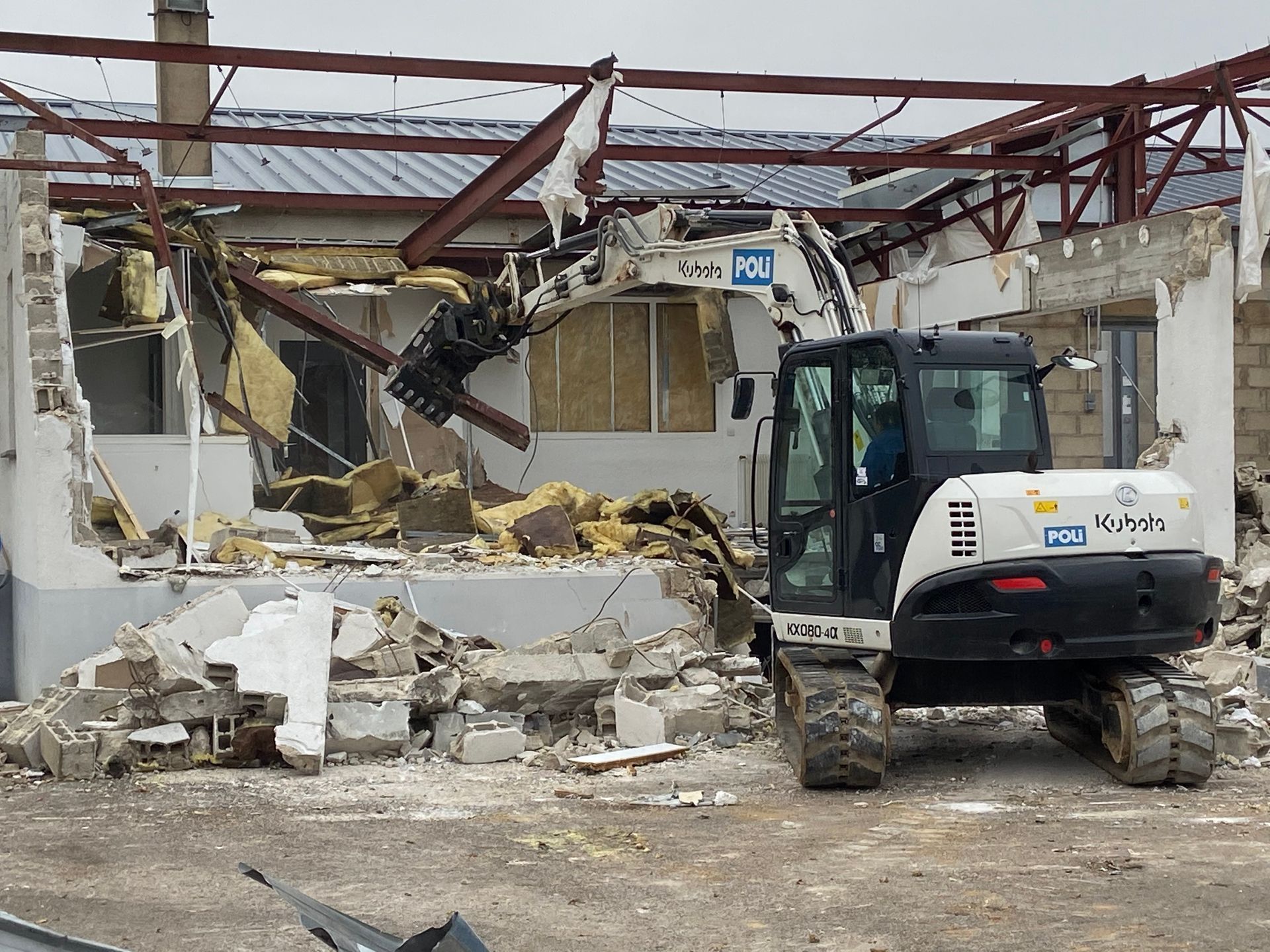 Chantier de démolition avec une pelleteuse en train de travailler sur des débris de construction. Des structures métalliques sont visibles parmi les débris.
