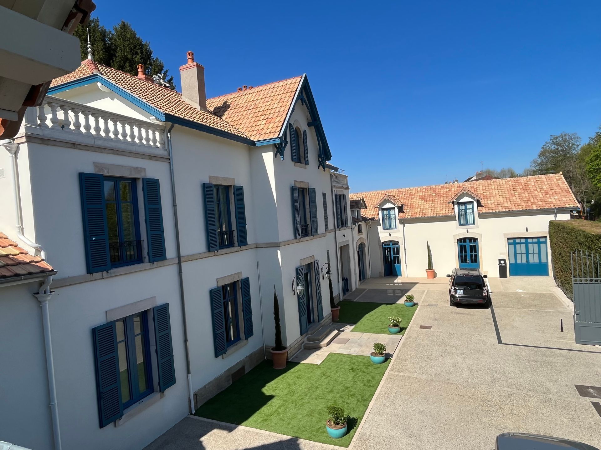 Maison moderne avec un jardin bien entretenu et une façade blanche. La maison est entourée de verdure et le ciel est clair et ensoleillé.