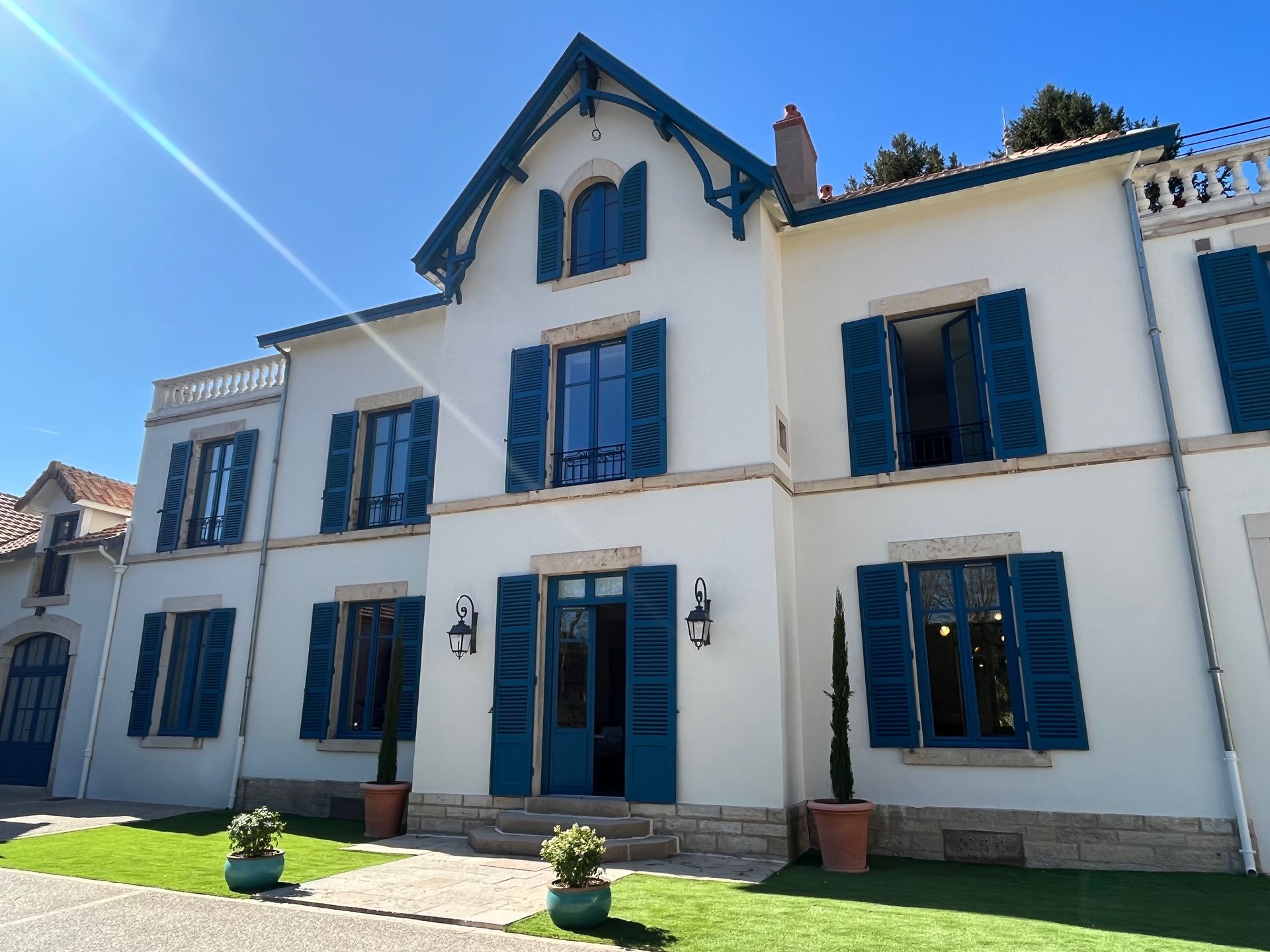 Maison moderne avec un jardin bien entretenu et une façade blanche. La maison est entourée de verdure et le ciel est clair et ensoleillé.