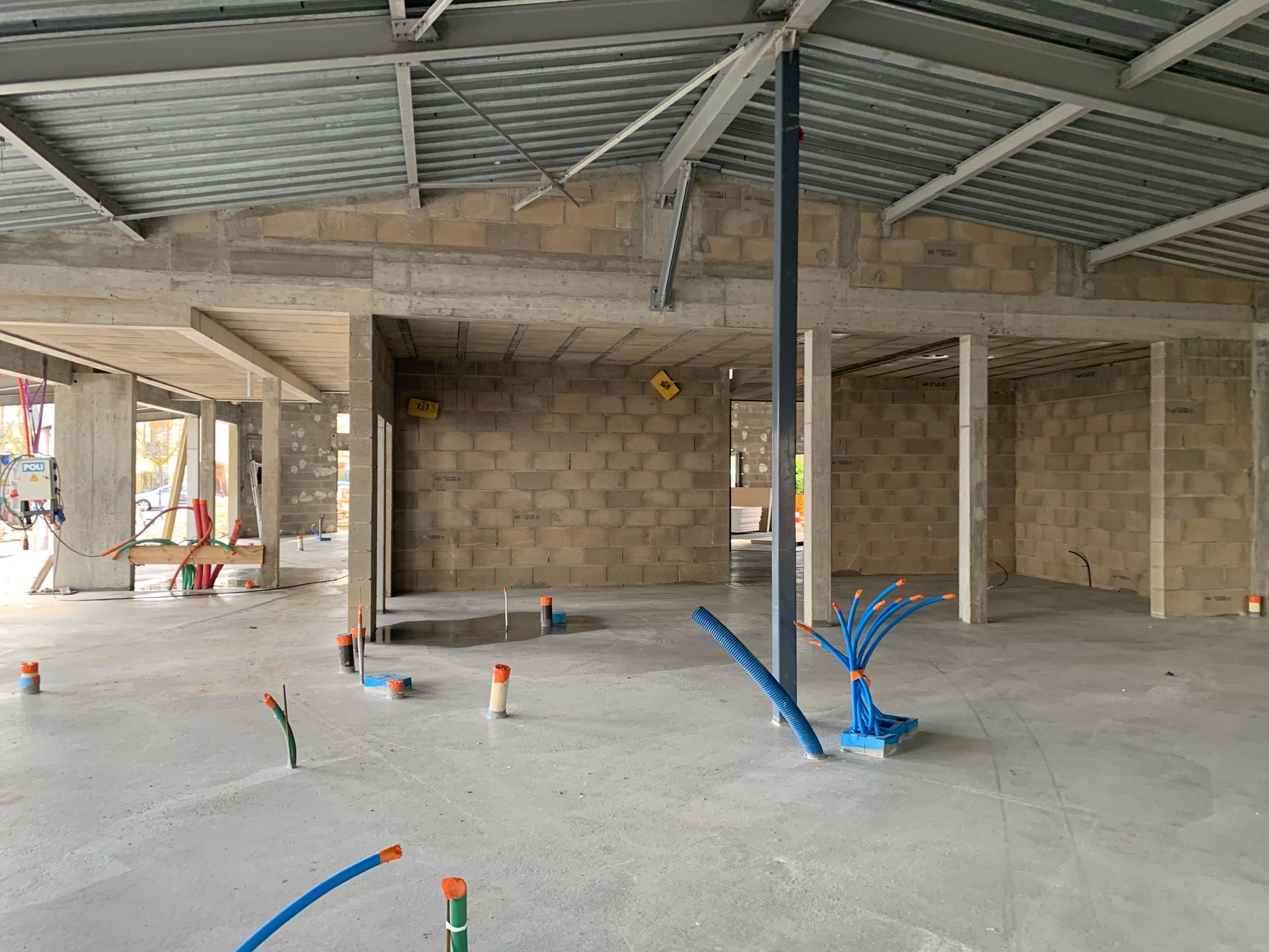 Intérieur d'un bâtiment en construction avec des structures en métal et des murs en béton. On peut voir des outils et des matériaux de construction éparpillés sur le sol.
